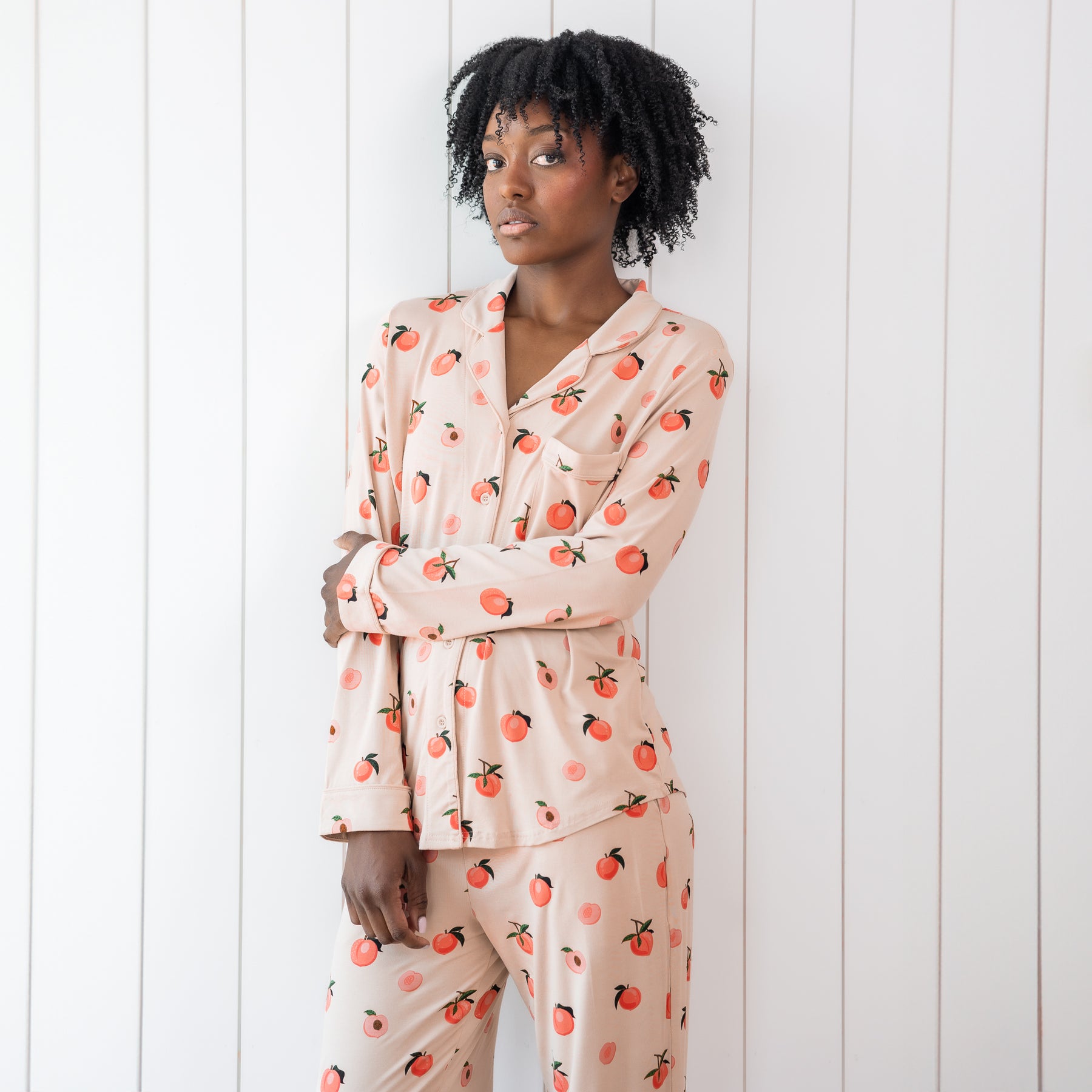 Woman modeling light pink button up bamboo pajamas in a peach print against white wooden wall
