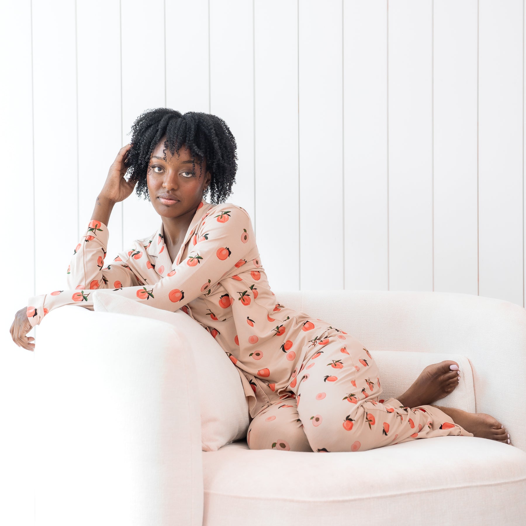 Woman modeling light pink button up bamboo pajamas in a peach print sitting in a cream chair with white wall backdrop