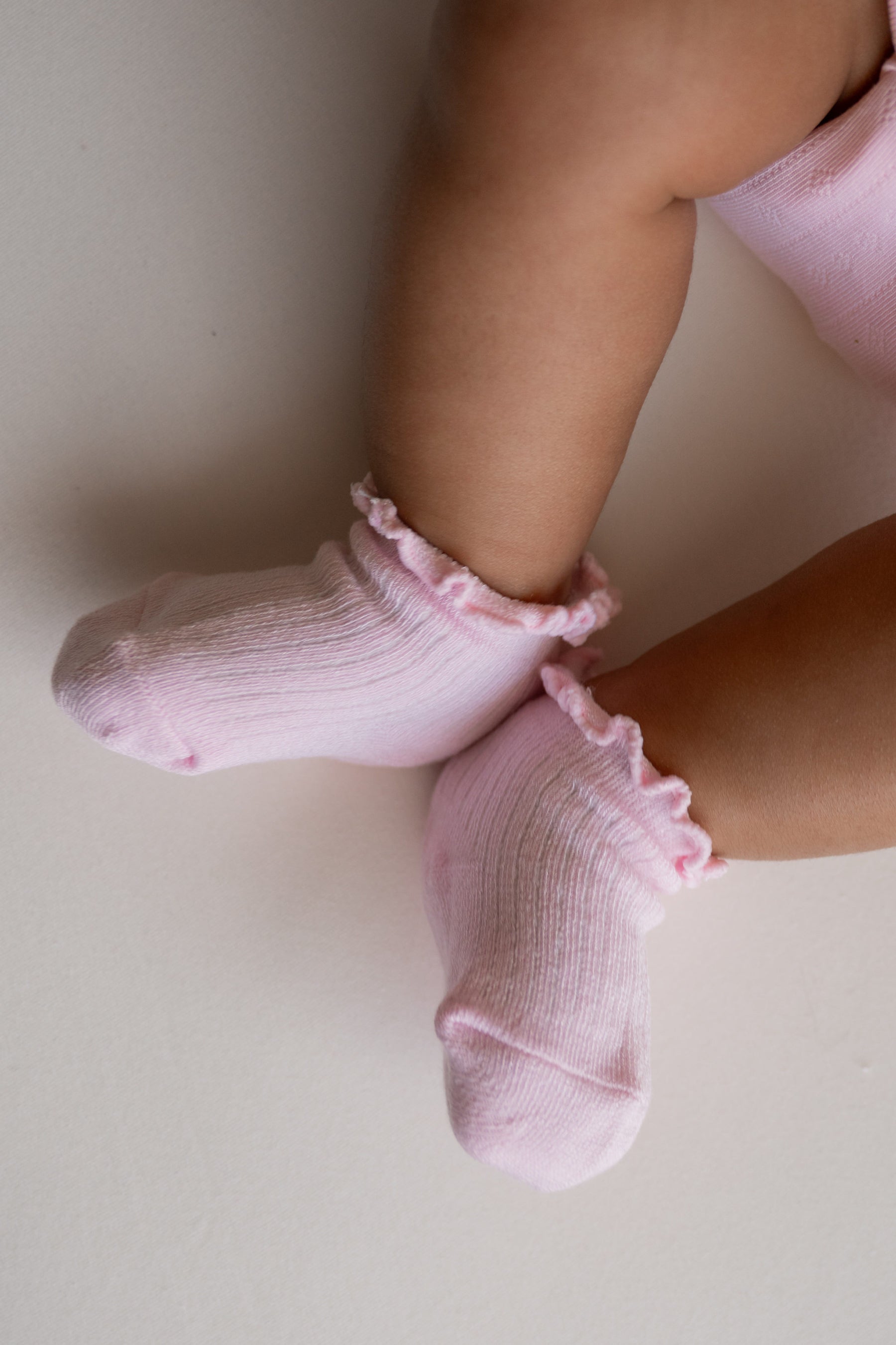A close-up of a baby's legs wearing light pink ribbed ankle socks with ruffled trim, resting on a light beige surface.