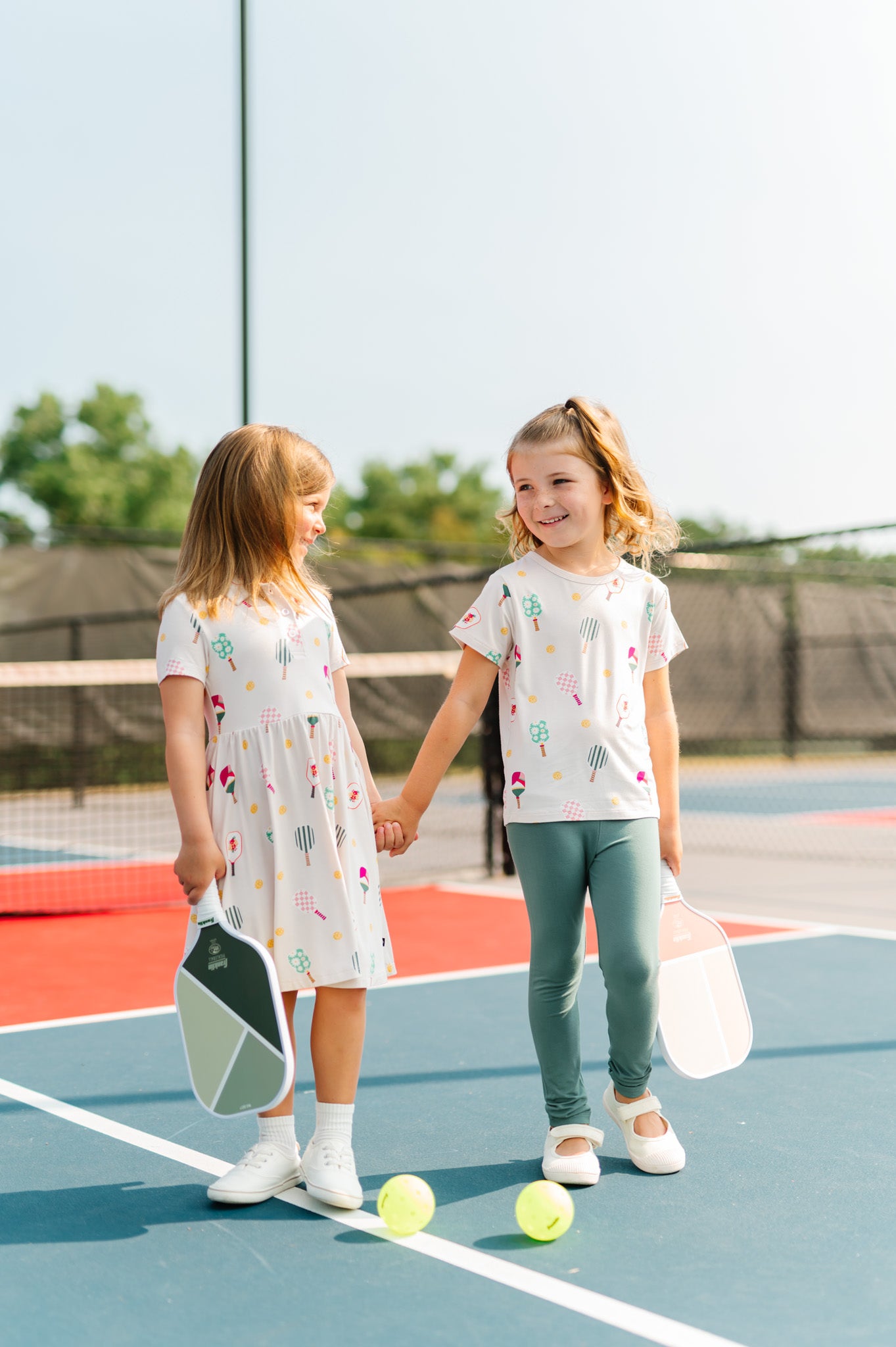 Two girls on pickleball court holding hands and bats wearing Polo Dress and Toddler Basic Tee in Pickleball print
