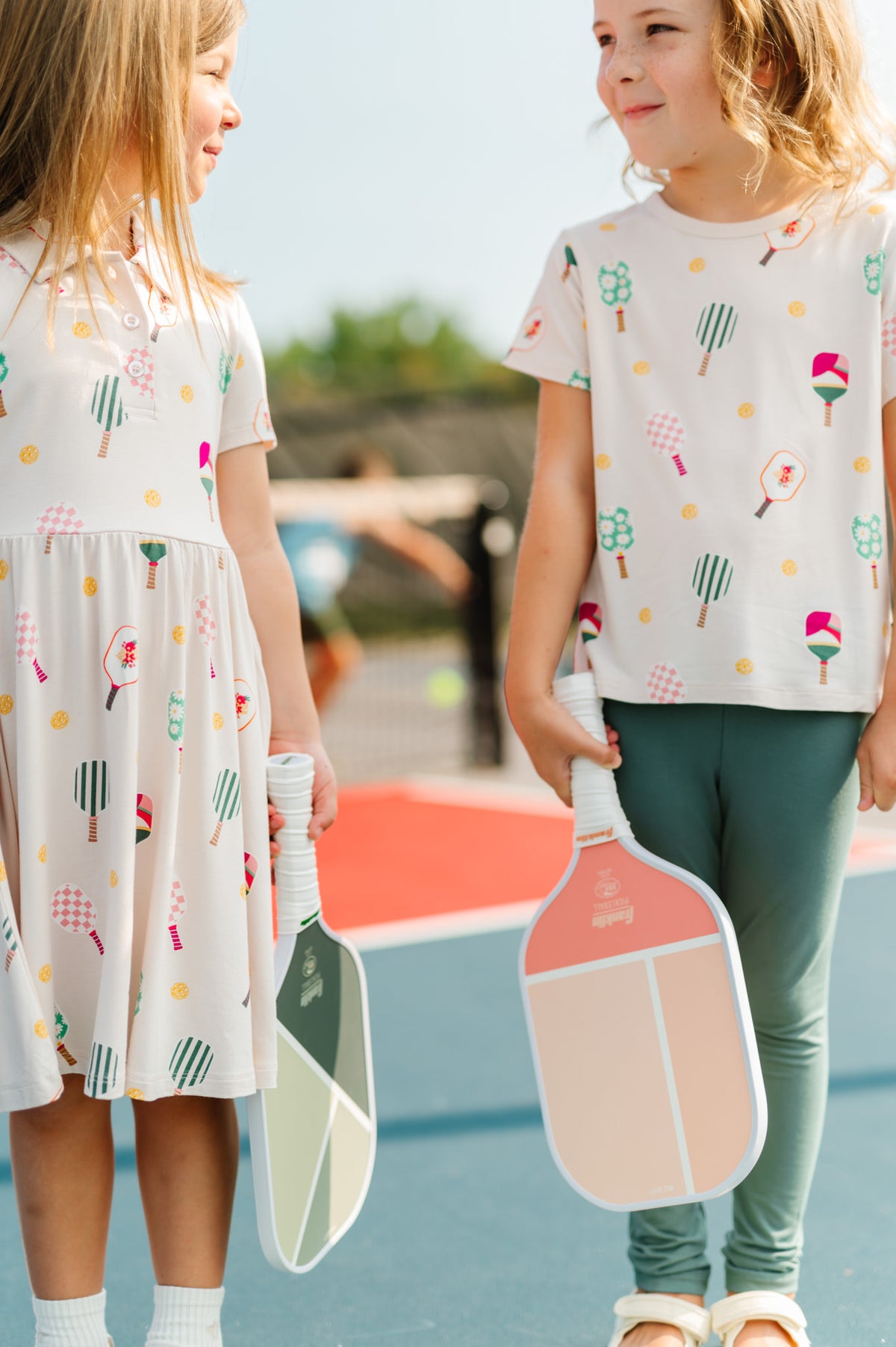 Two girls on pickleball court holding bats wearing Polo Dress and Toddler Basic Tee in Pickleball print
