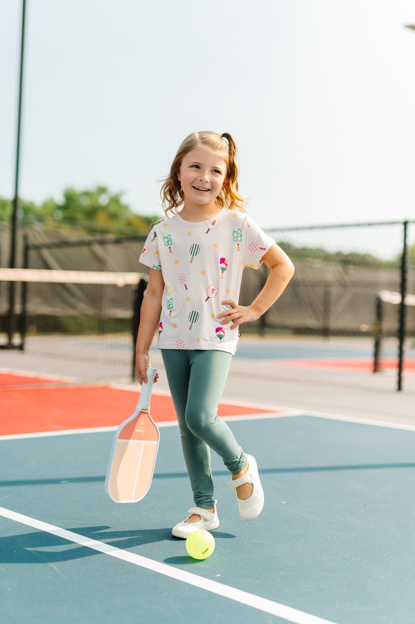 Girl posing in Toddler Basic Tee in Pickleball print over green leggings  holding a bat on pickleball court