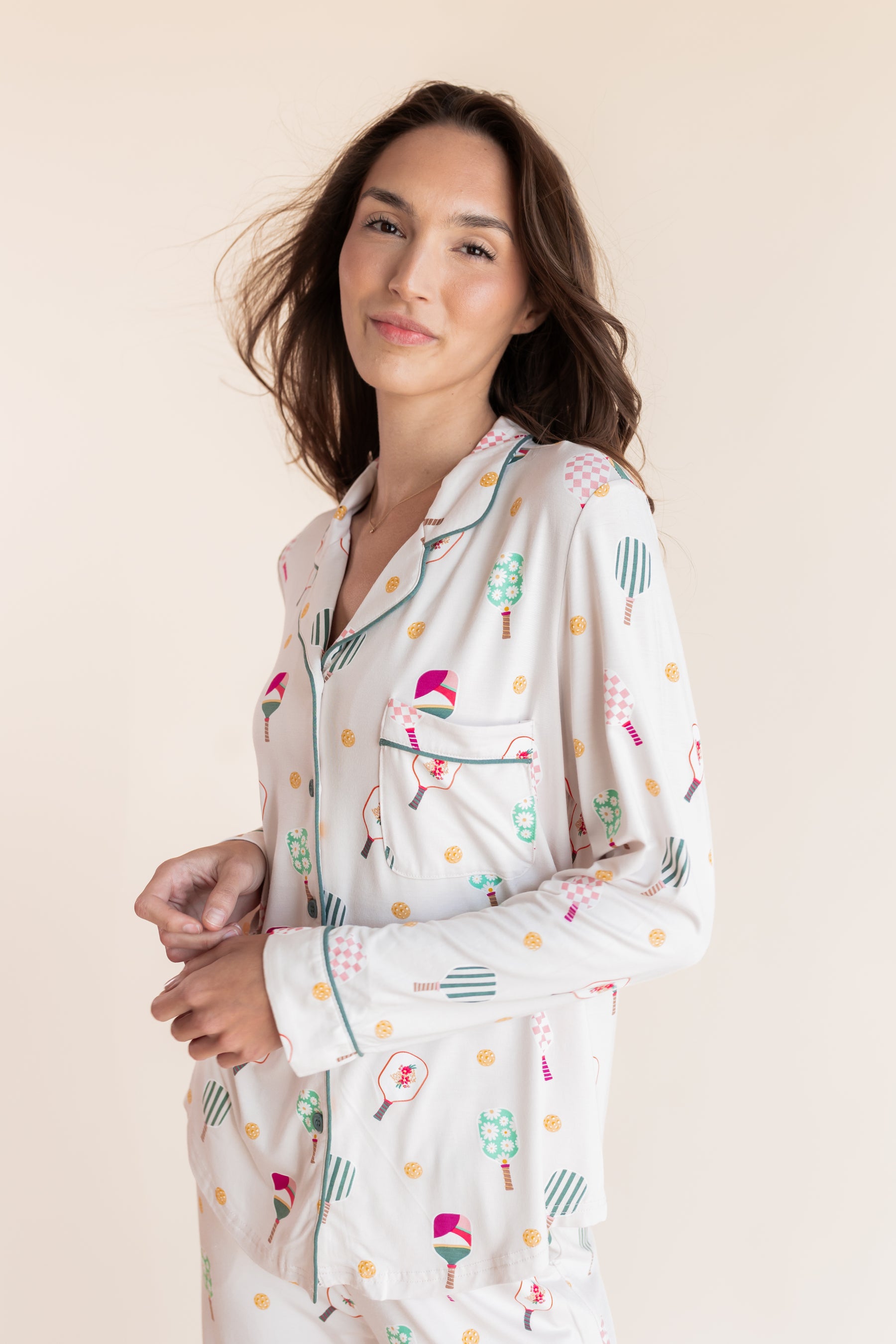 A woman wearing a long-sleeve pajama shirt with a colorful pickleball pattern on a cream background, posed against a solid light beige backdrop.