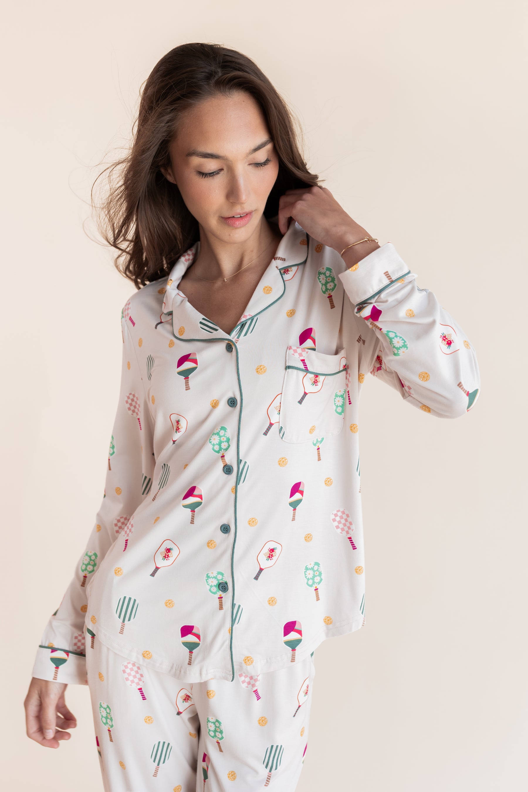 A woman wearing a long-sleeve button-front pajama top and matching pajama pants with a whimsical multicolor print of pickleball rackets on a cream background, set against a solid light beige studio backdrop.