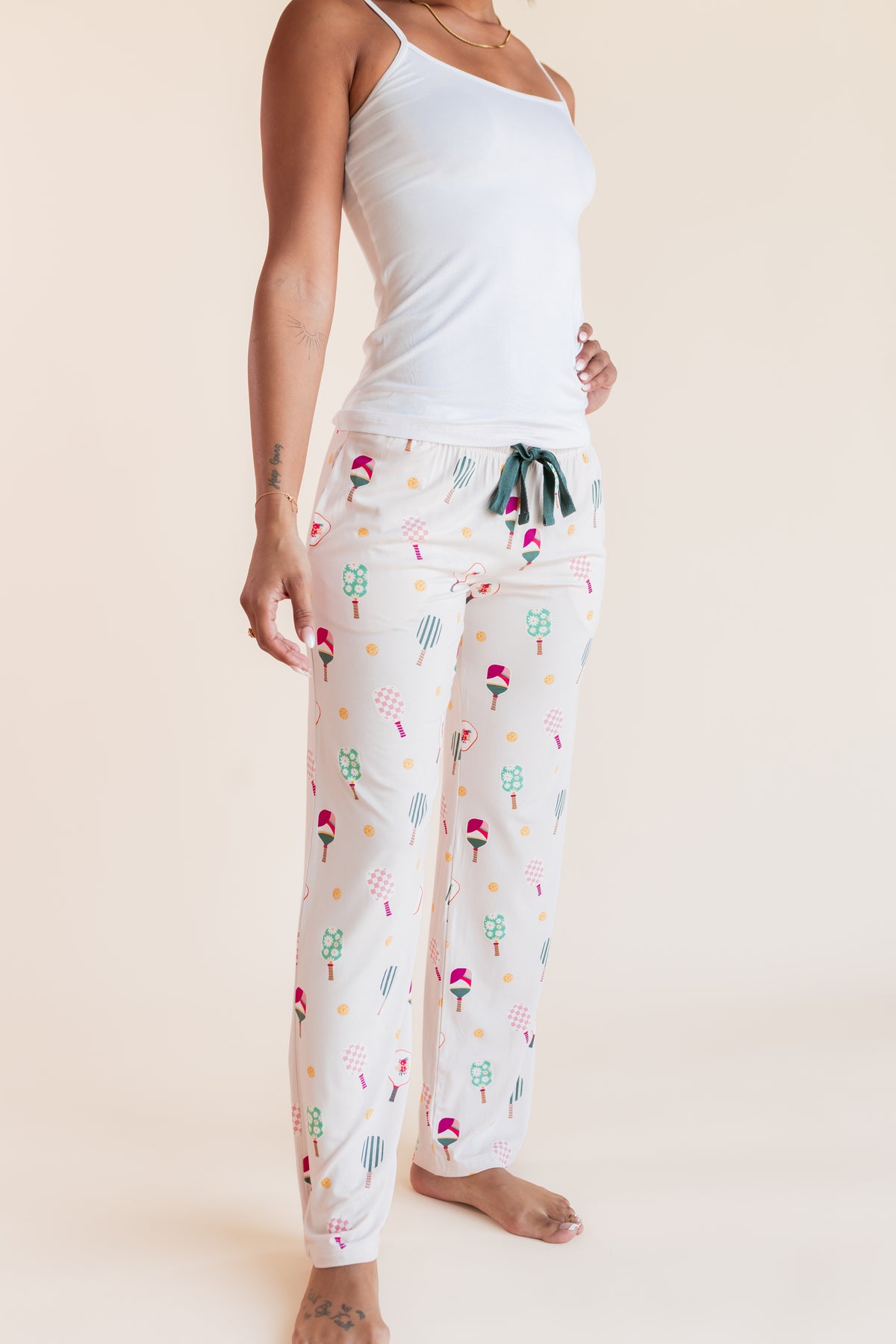 A woman modeling white pajama pants with a colorful pickleball pattern, paired with a white tank top, against a solid beige background.