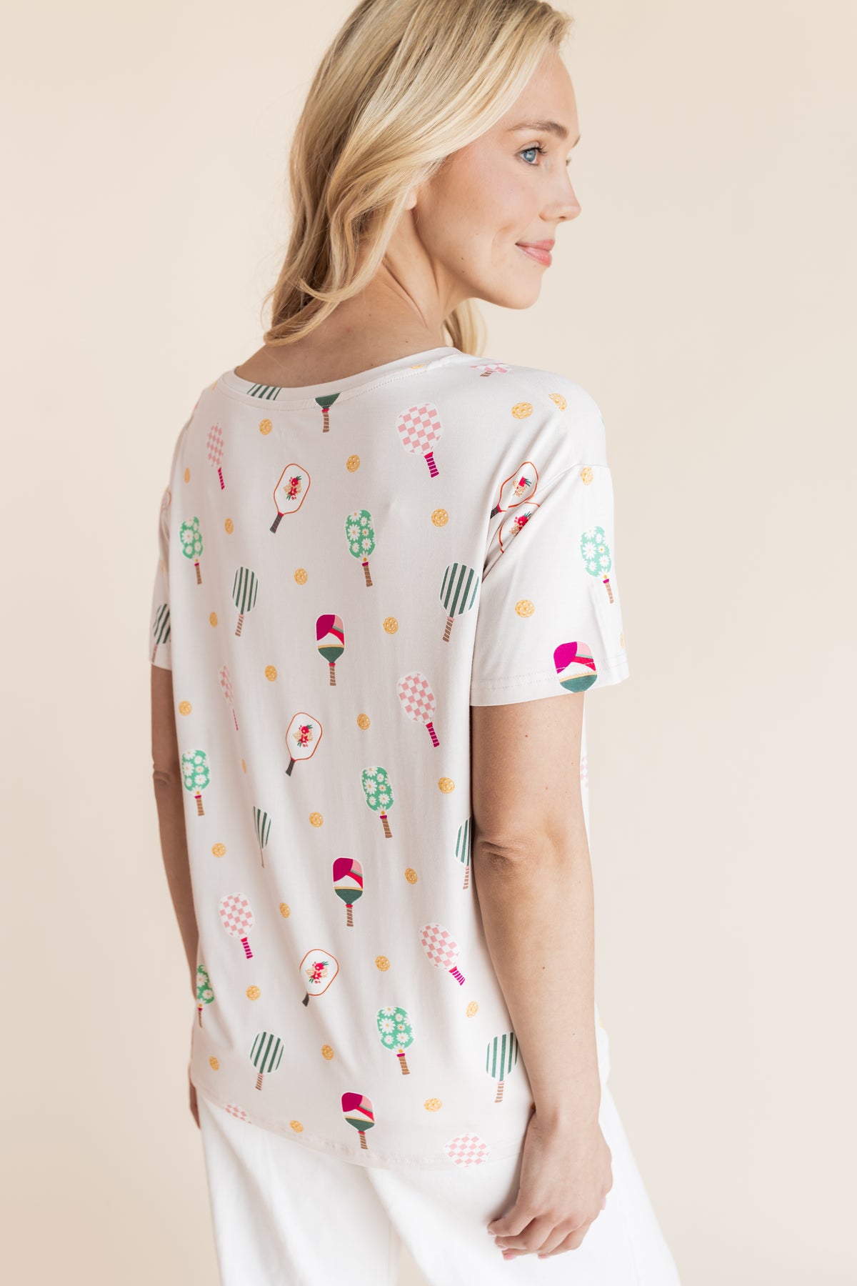 A woman shown from the back wearing a white short-sleeve t-shirt with a colorful pickleball print paired with white pants, against a solid beige background.