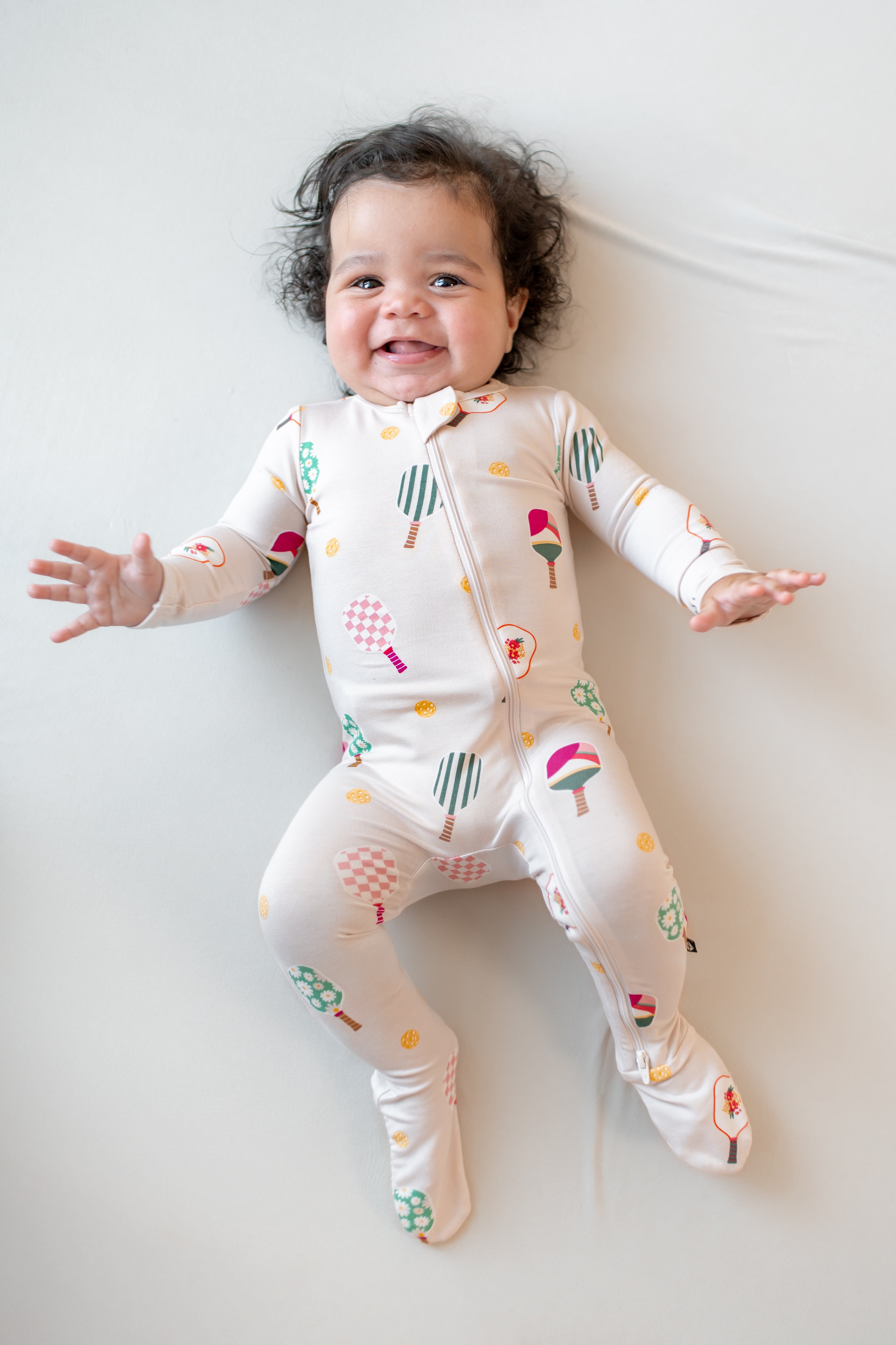 Baby wearing Zippered Footie in Pickleball print on a beige background