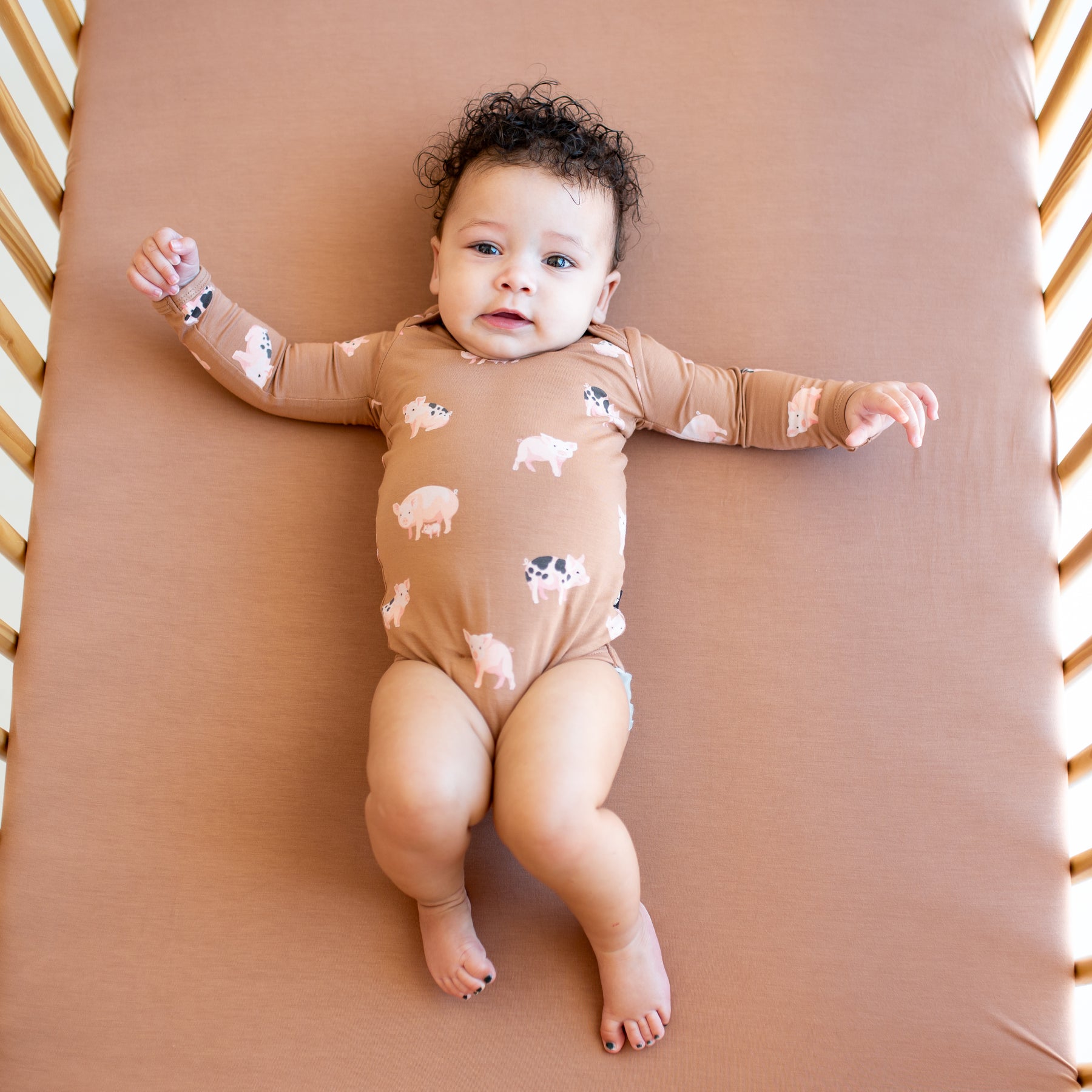 Infant laying in a crib on a Latte crib sheet wearing the Long Sleeve Bodysuit in Pig