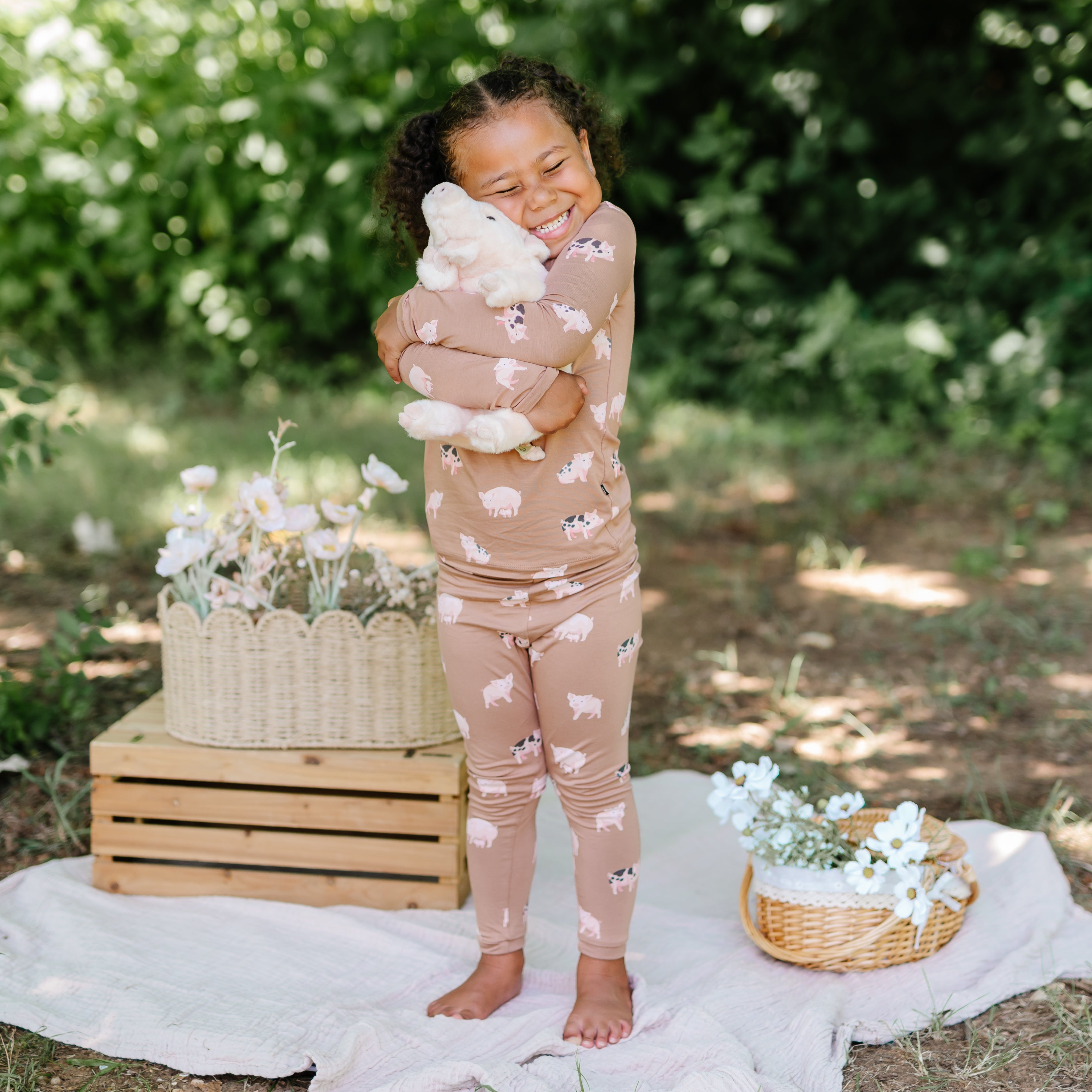 Smiling girl hugging a stuffed toy big outside standing on a white blanket with flowers in wicker baskets on either side of her wearing the Long sleeve toddler pajamas in Pig