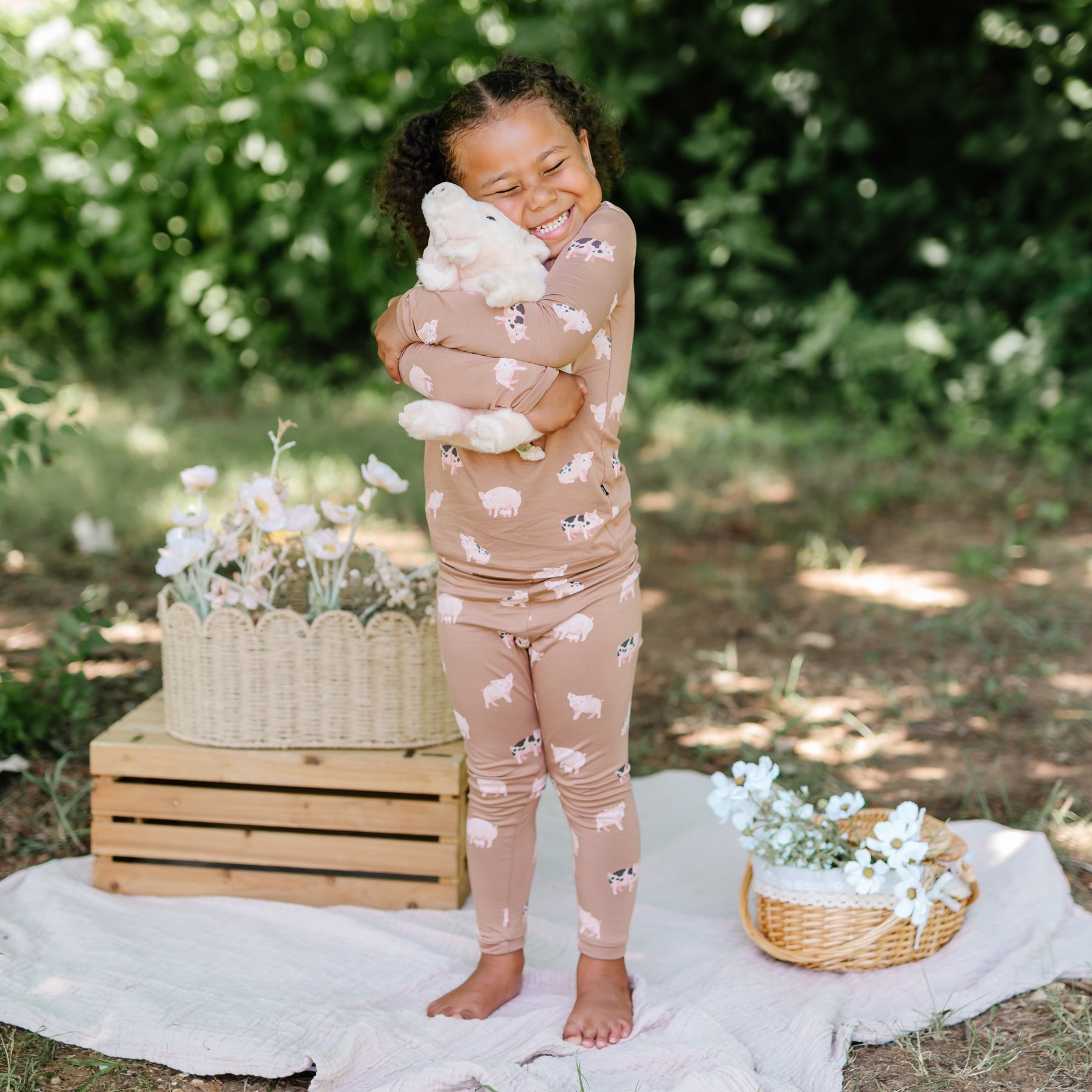 Smiling girl hugging a stuffed toy big outside standing on a white blanket with flowers in wicker baskets on either side of her wearing the Long sleeve toddler pajamas in Pig