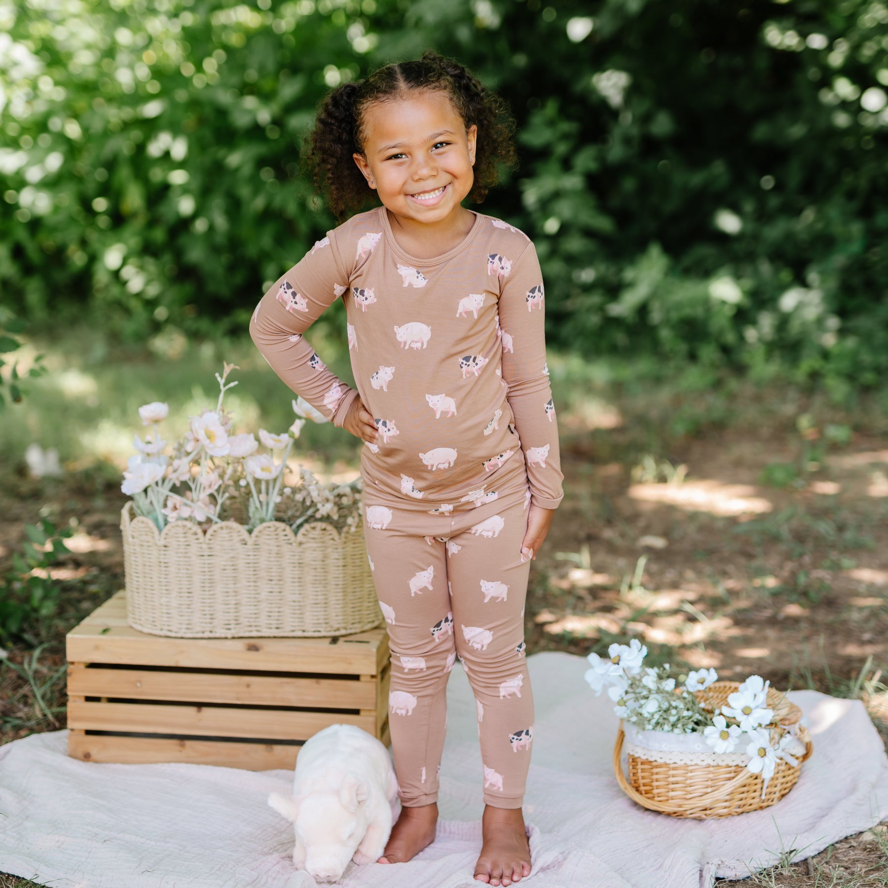 Young girl standing outside with one hand on hip between flowers in wicker baskets, beside a stuffed toy pig on a white blanket wearing the Long Sleeve Pajamas in Pig