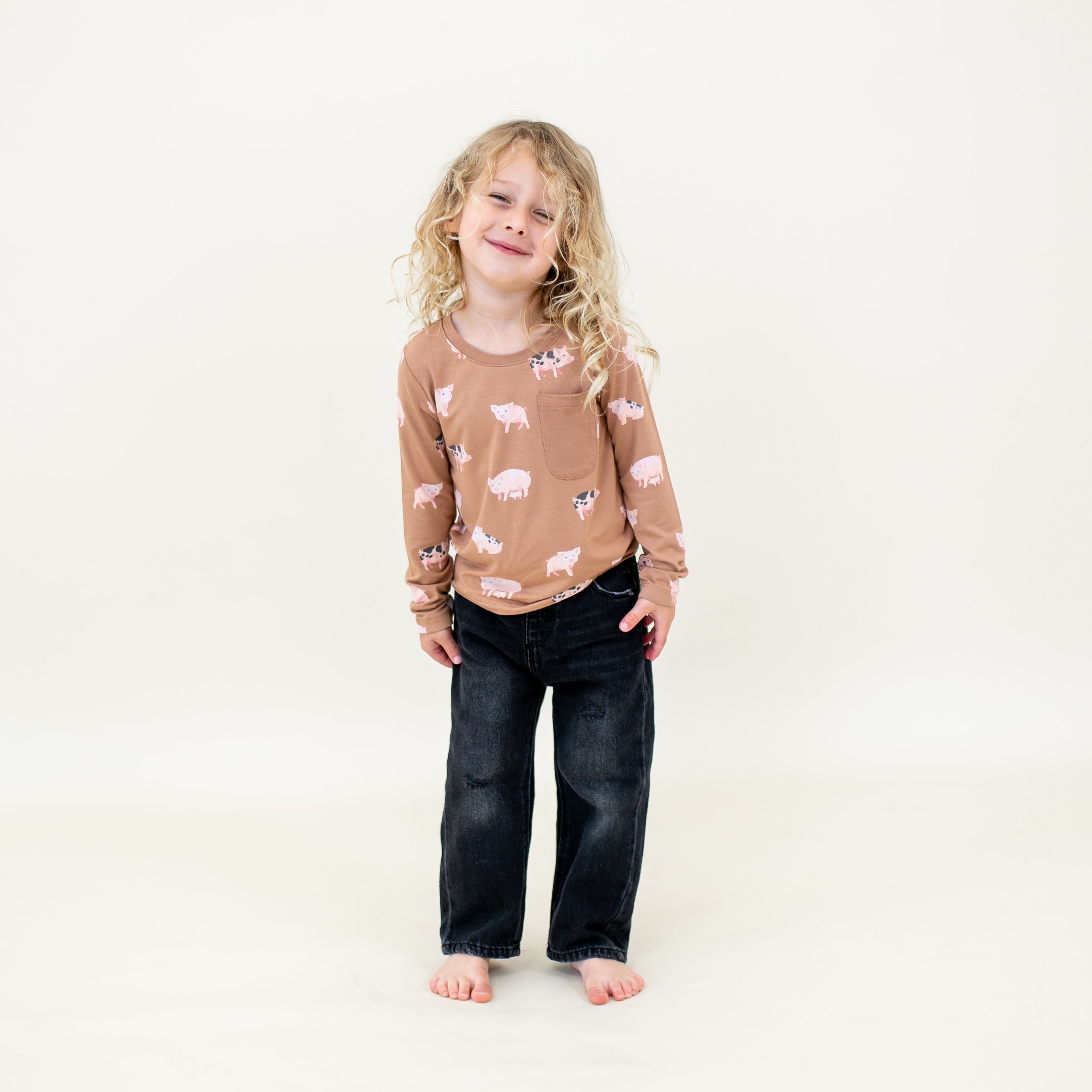Smiling child wearing the Long Sleeve Toddler Crew Neck Tee in Pig with black jeans standing in front of a light neutral background