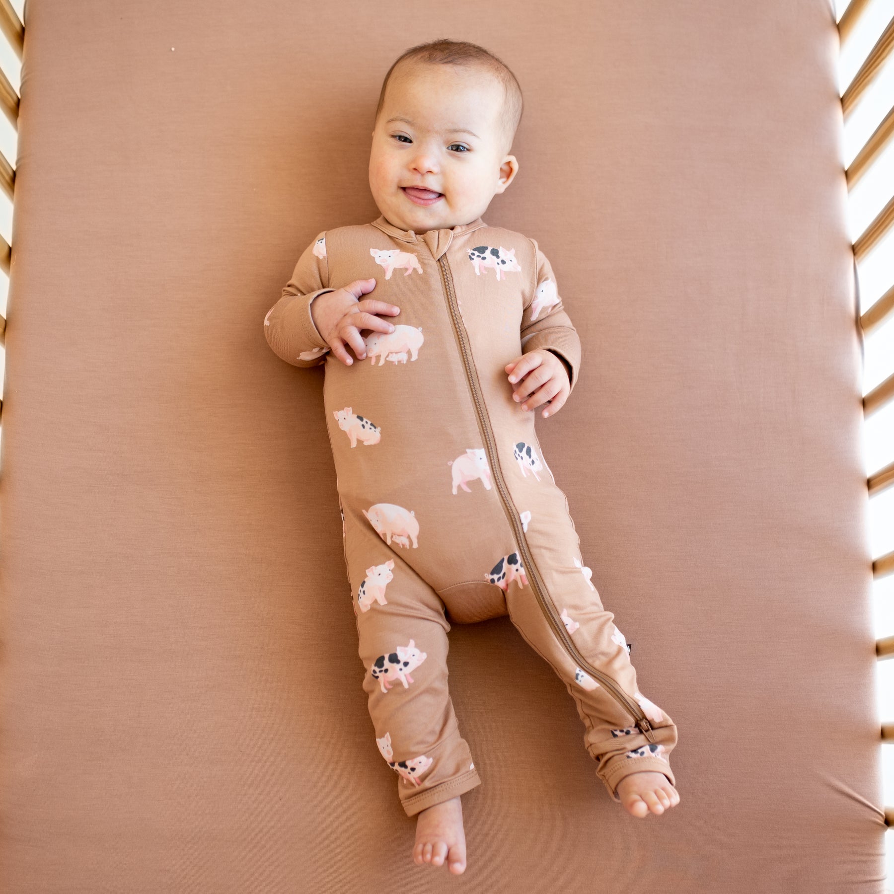 Infant sticking her tongue out  laying in a crib on top of a latte crib sheet wearing the Zippered Romper in Pig