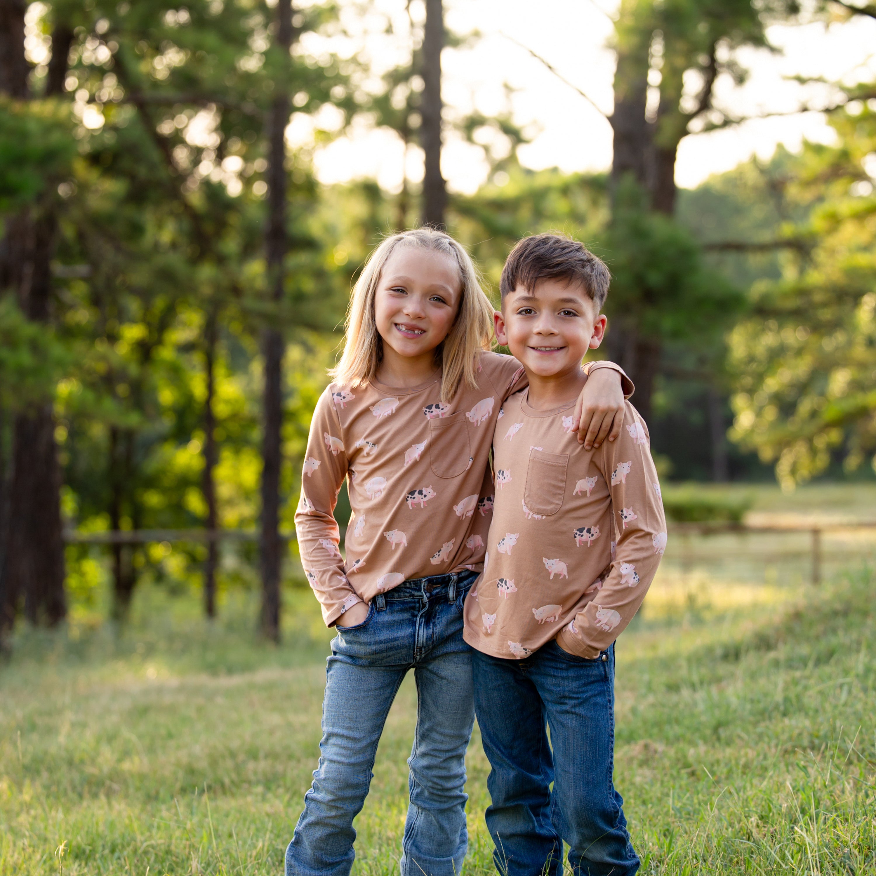 Brother and sister standing in a field with arms around one another shoulders wearing the Long Sleeve Toddler Crew Neck Tee in Pig paired with jeans
