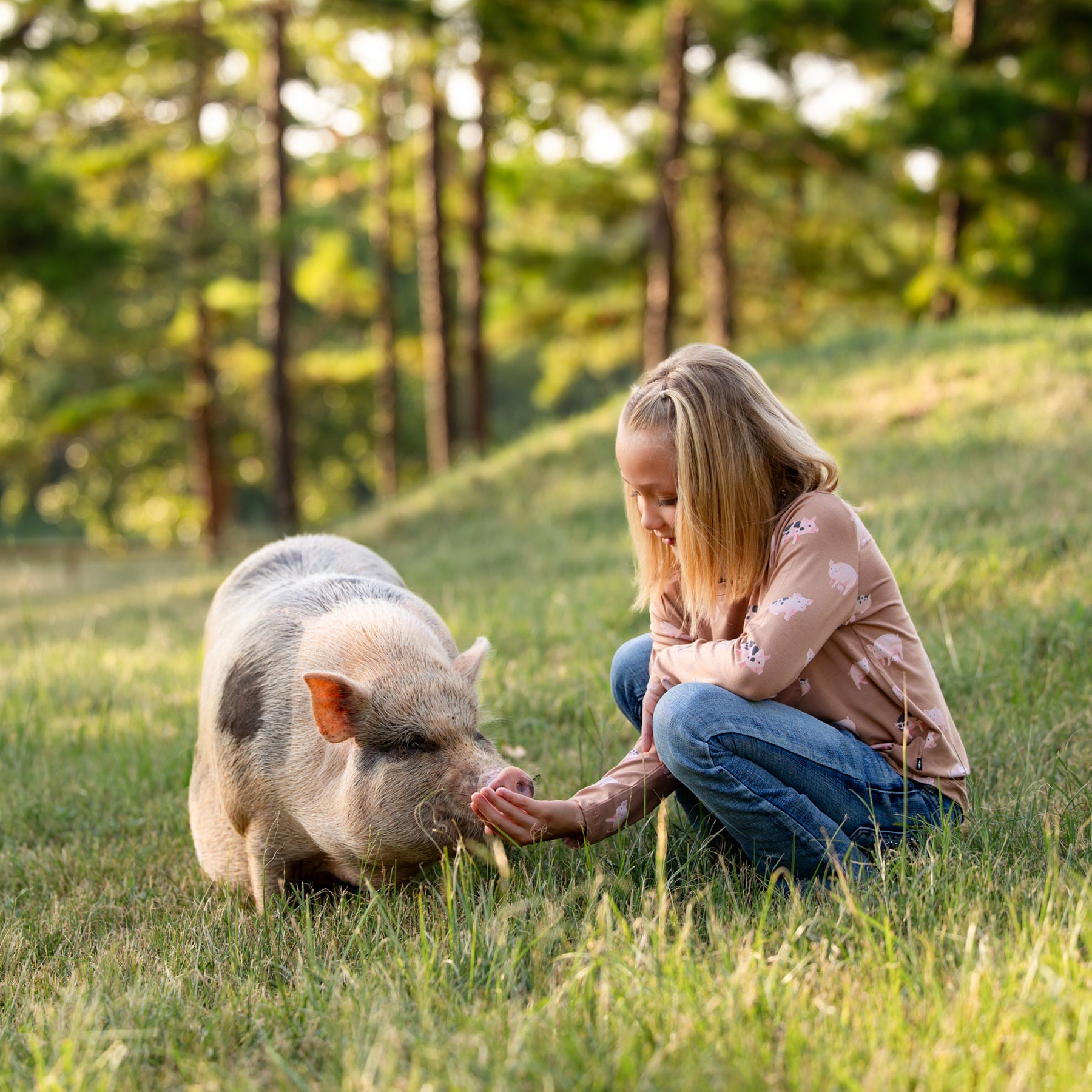 Girl outside crouched in the grass feeding her pet pig wearing the Long Sleeve Toddler Crew Neck Tee in Pig and jeans