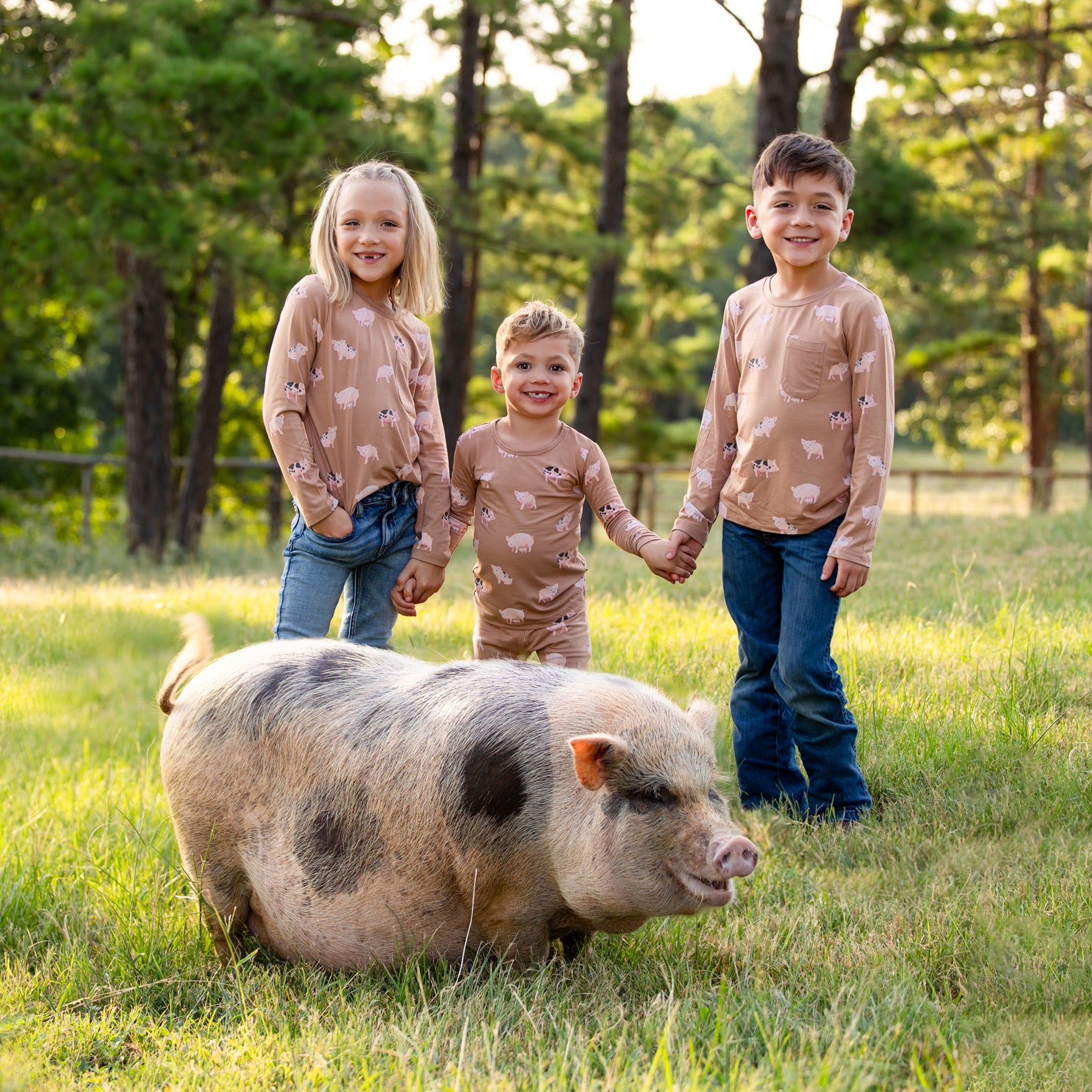 Brothers and sister holding hands standing in front of a field standing behind their pet pig wearing the Long Sleeve Pajamas in Pig and Long Sleeve Toddler Crew Neck in Pig