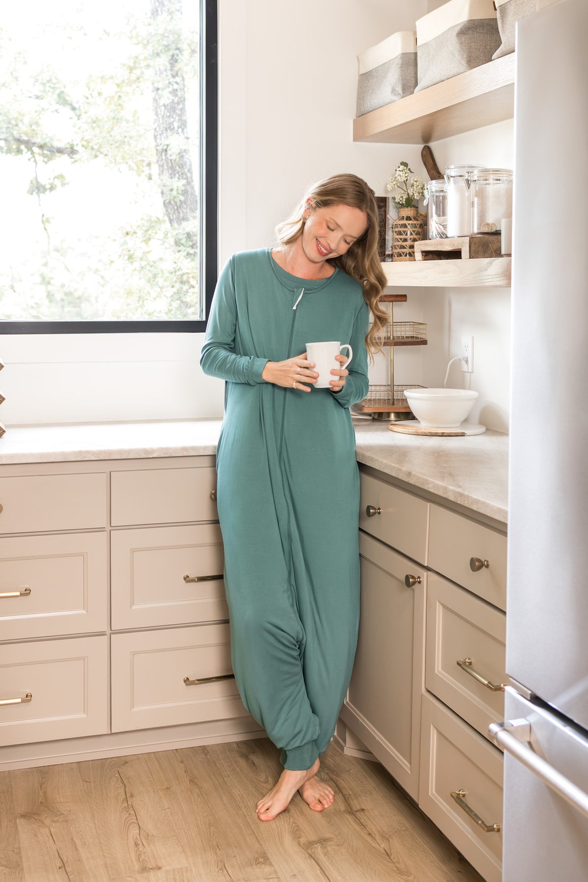 A woman wearing a sage green sleep sack stands barefoot in a bright kitchen, holding a white mug.