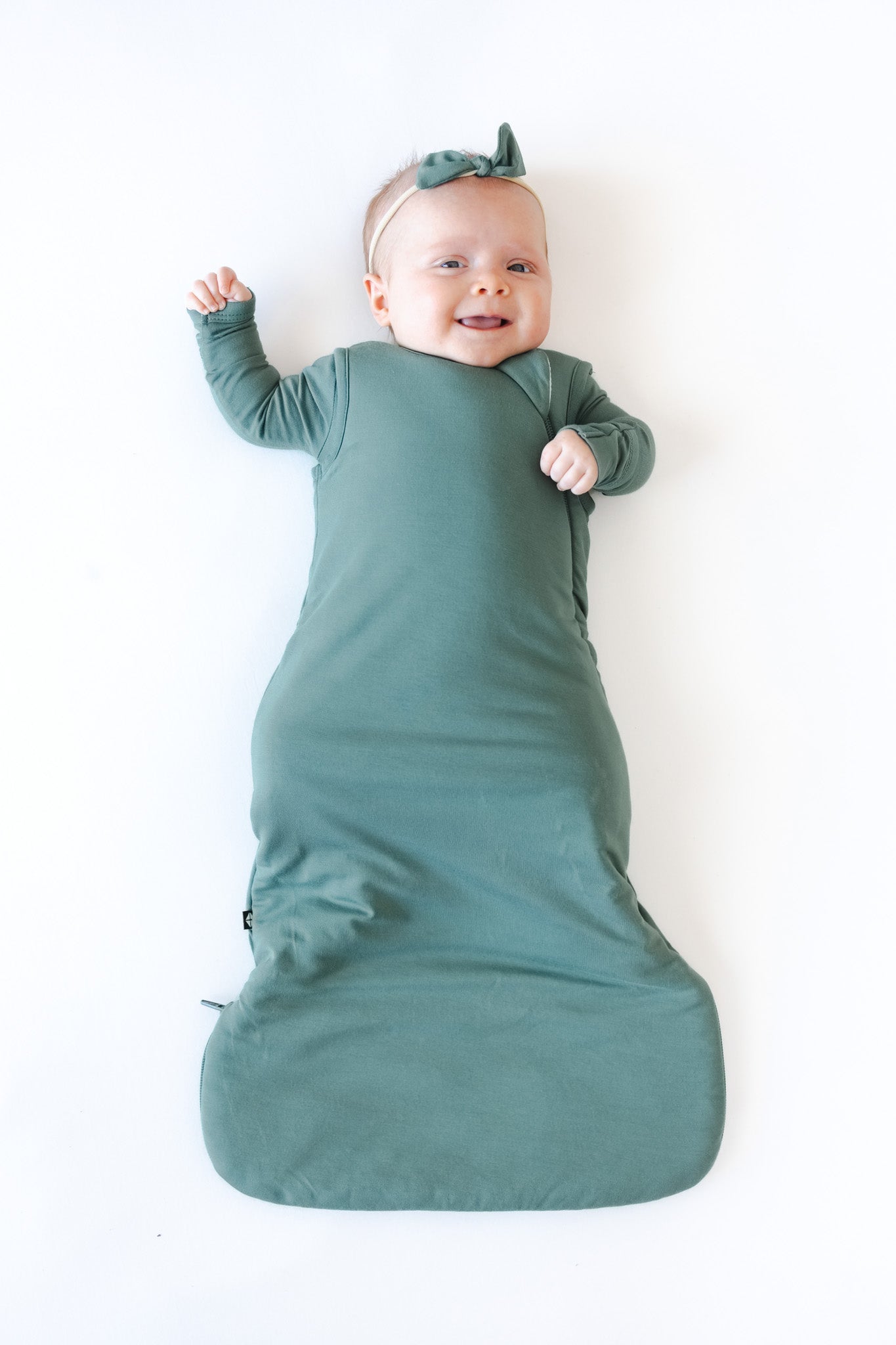 A baby lying on their back wrapped in a sage green sleep bag, wearing a matching green bow headband, against a solid white background.