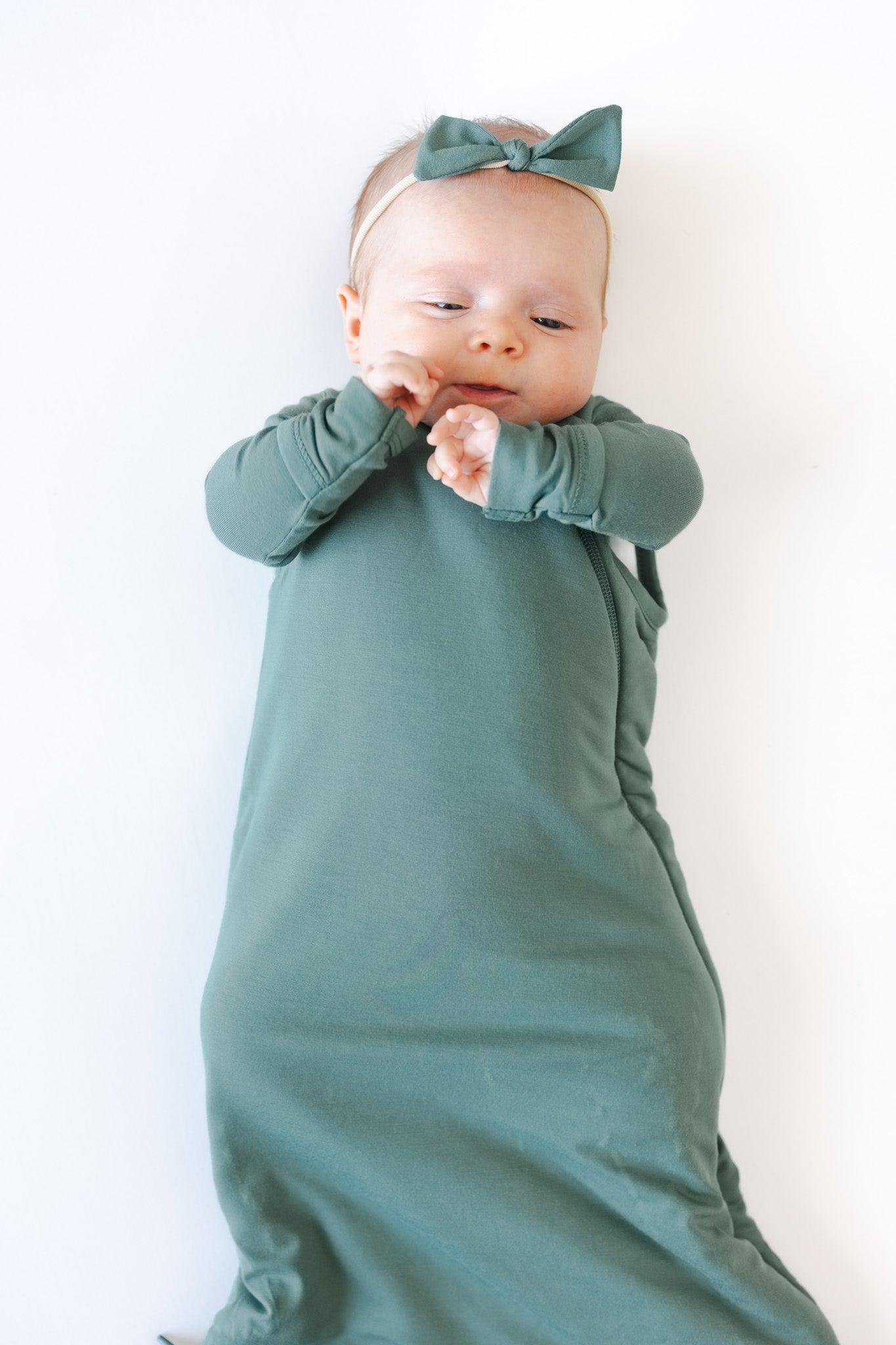 A baby wearing a sage green sleep bag and a matching green bow headband, lying on a plain white background.