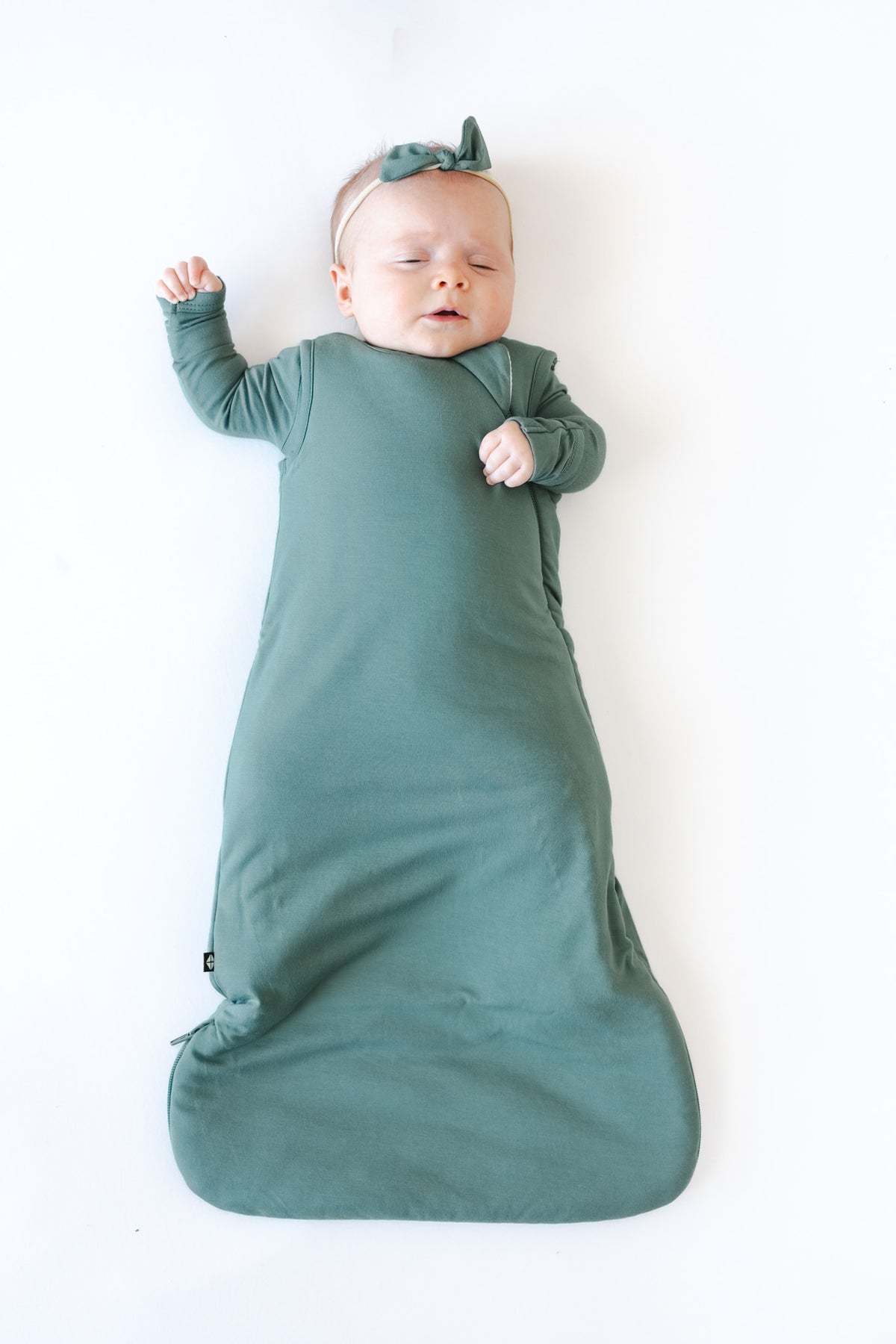 A sleeping newborn baby swaddled in a  sage green sleep sack, wearing a matching sage green bow headband, lying on a clean white background.