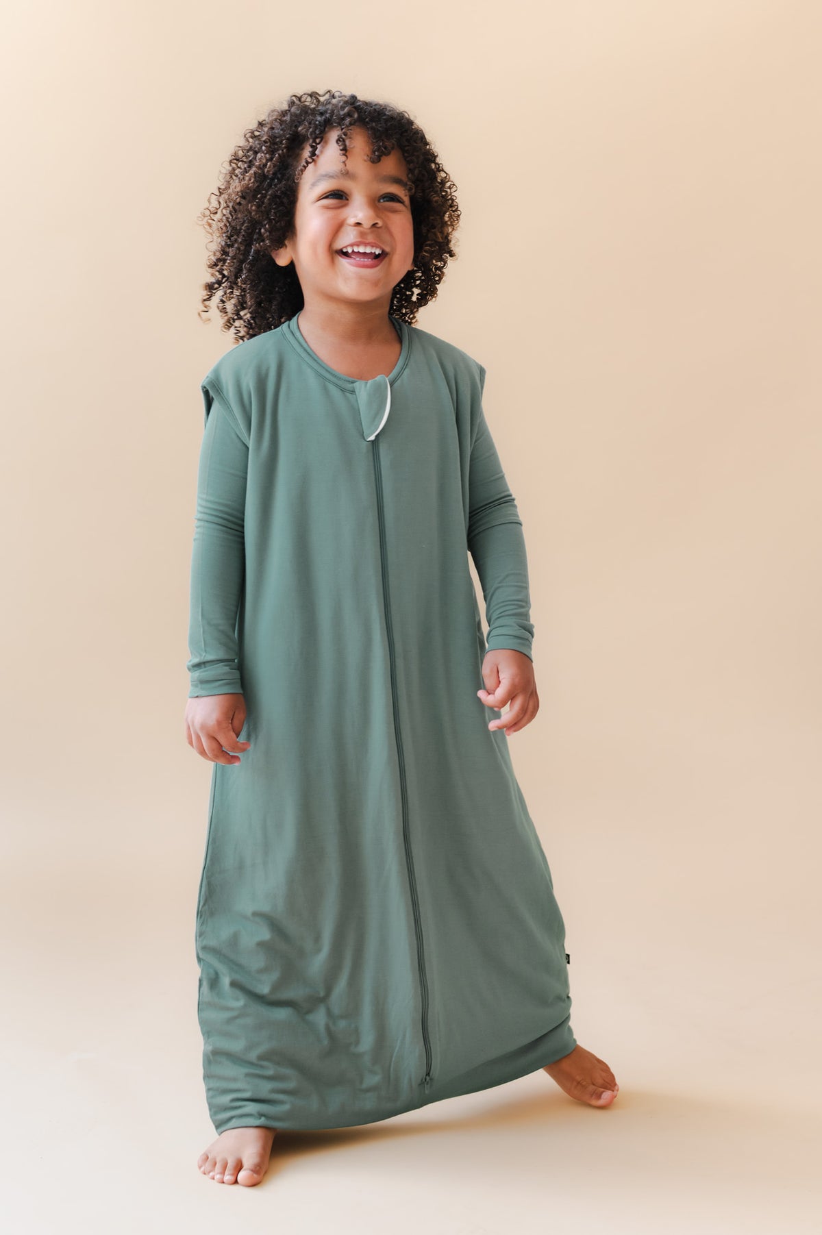 A smiling toddler wearing a sage green footless sleep sack, standing barefoot against a solid beige studio background.