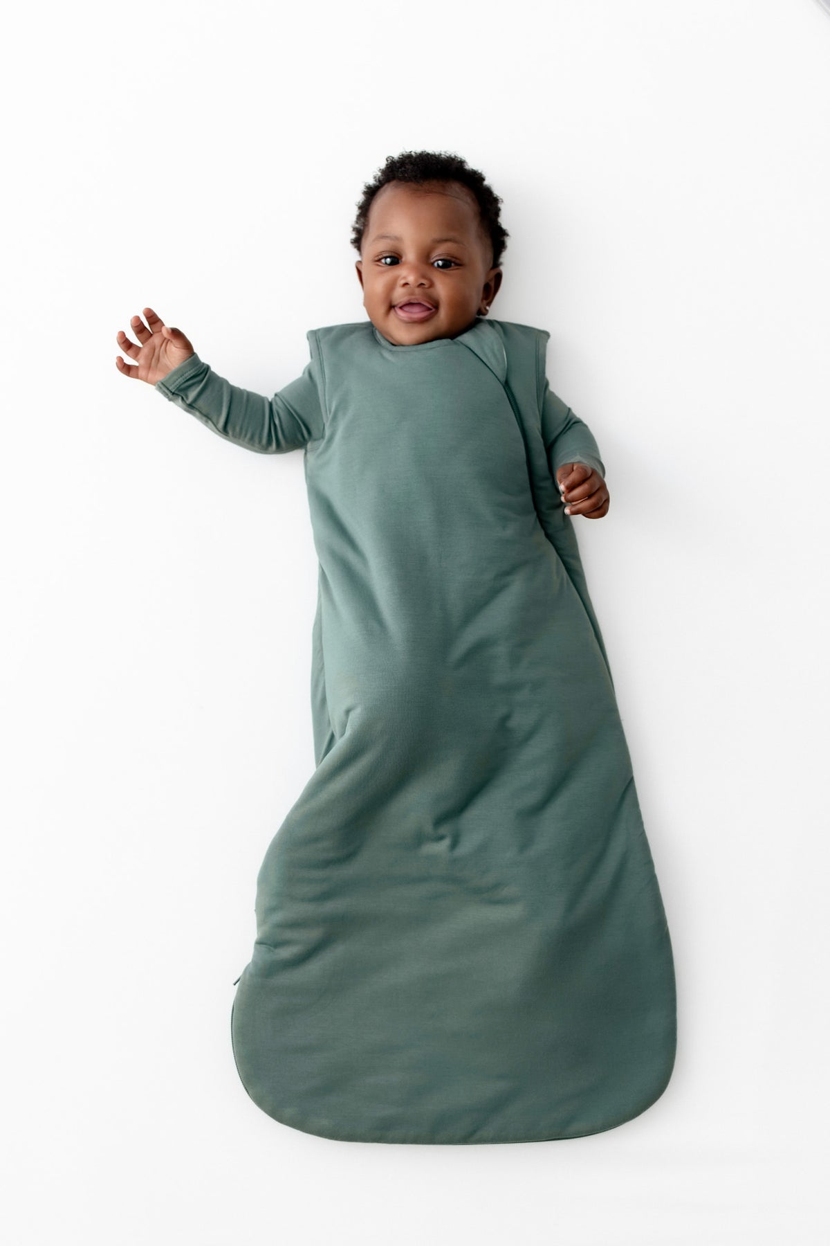 A baby wearing a sage green 2.5 TOG sleep sack, lying on their back against a clean white background.