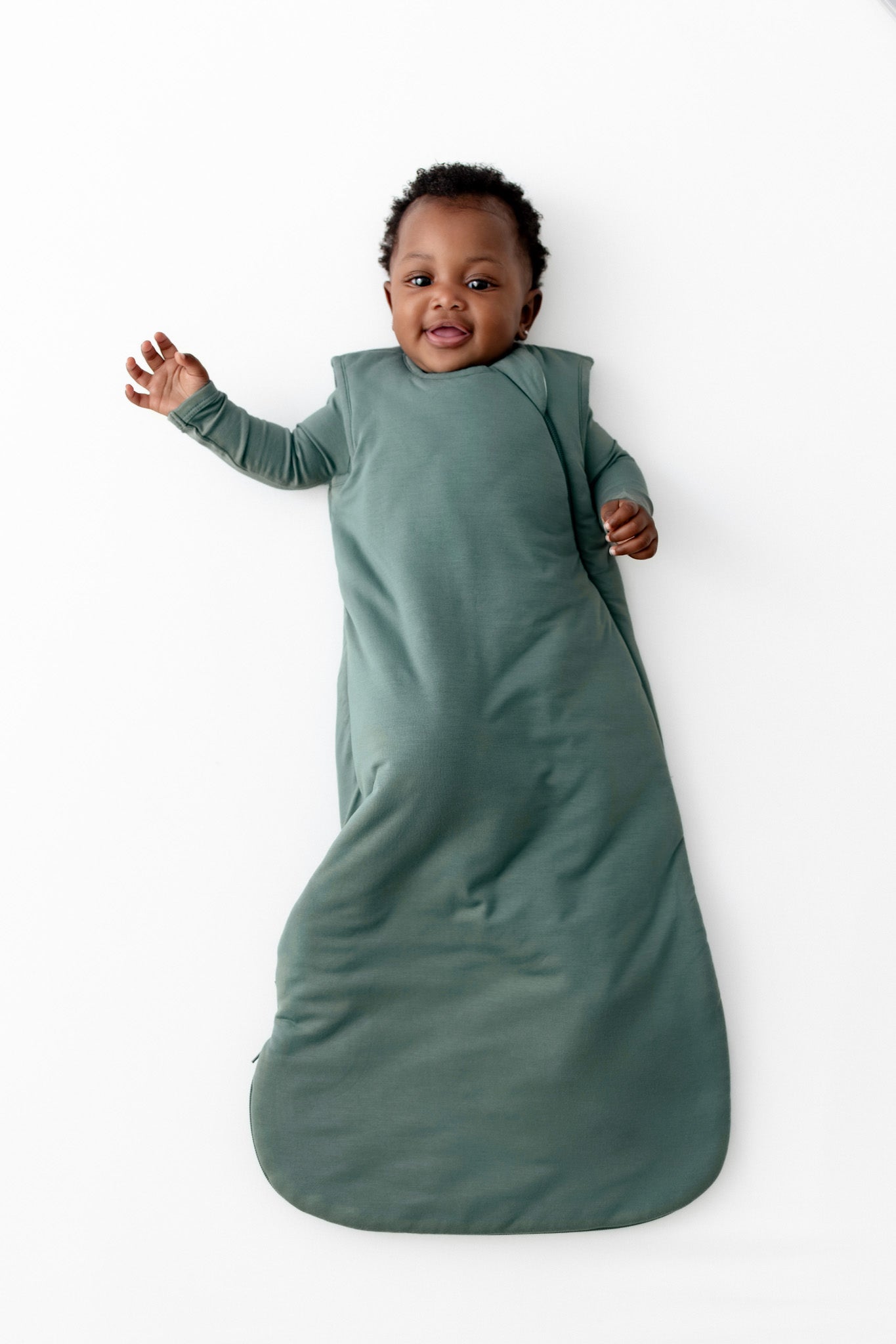 A baby wearing a sage green 2.5 TOG sleep sack, lying on their back against a clean white background.