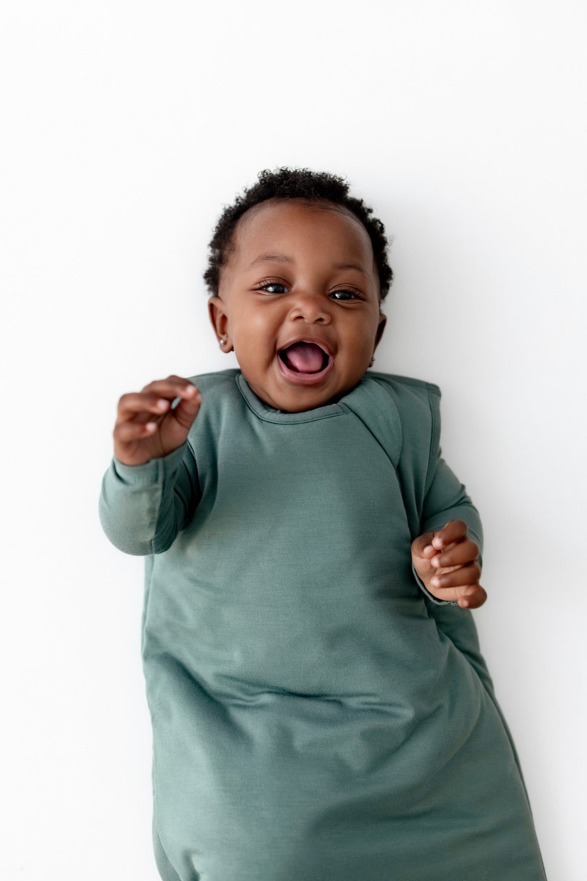 A smiling baby lying on their back against a plain white background, wearing a sage green sleep sack.