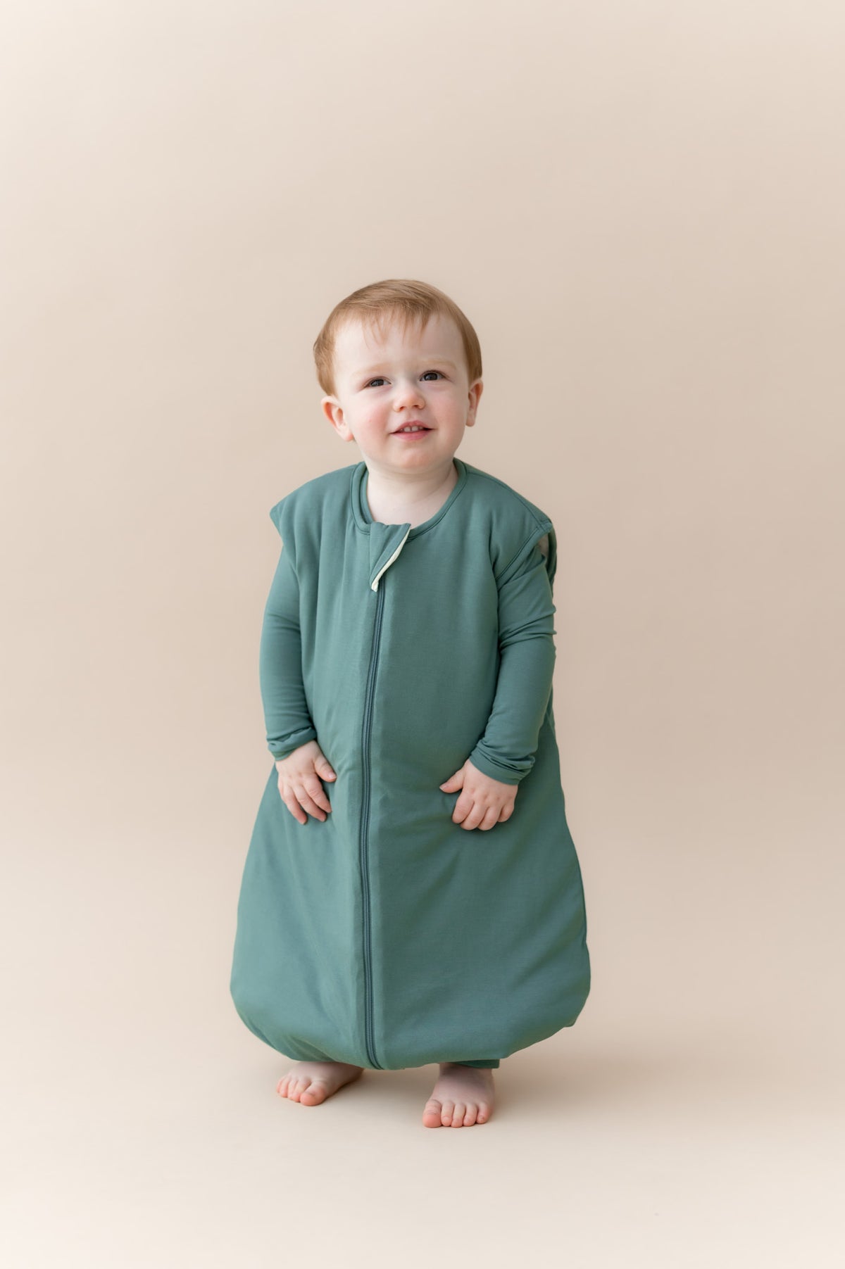 A toddler standing and wearing a sage green zippered sleep sack against a solid beige studio background.
