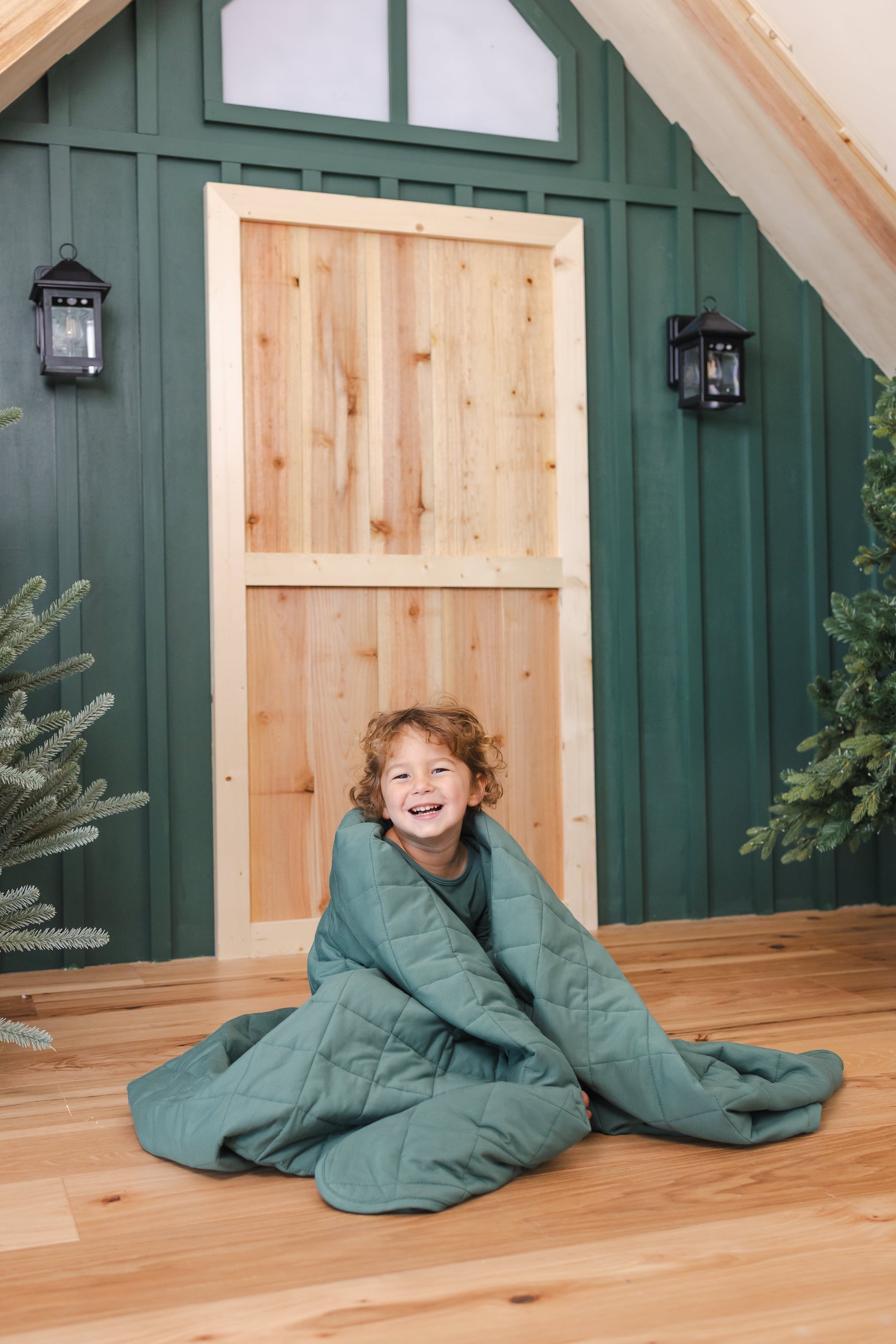 Toddler wrapped in Youth Blanket in Pine 2.5 sitting in front of  door