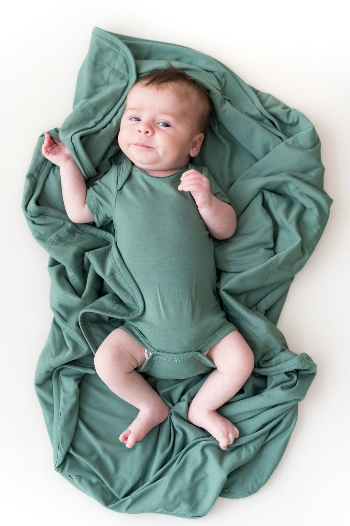 Baby lying on Baby Blanket in Pine wearing a similar color bodysuit on a beige background