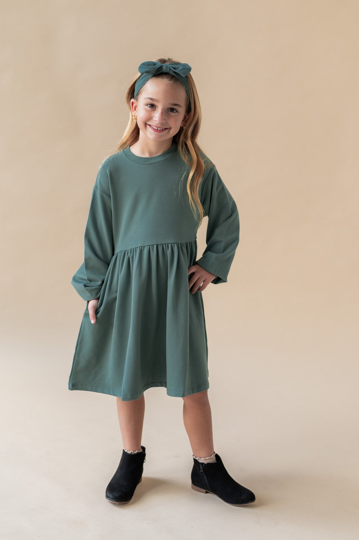 Girl smiling in Long Sleeve Skater Dress in Pine color with similar color knotted bow headband on a beige background