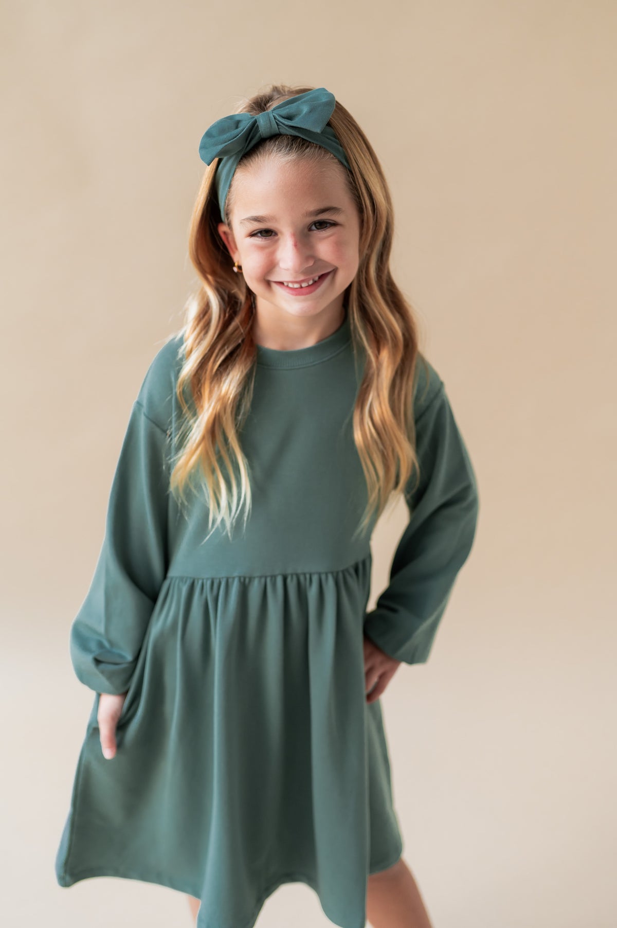 Girl wearing Long Sleeve Skater Dress in Pine color with similar color knotted bow headband on a beige background