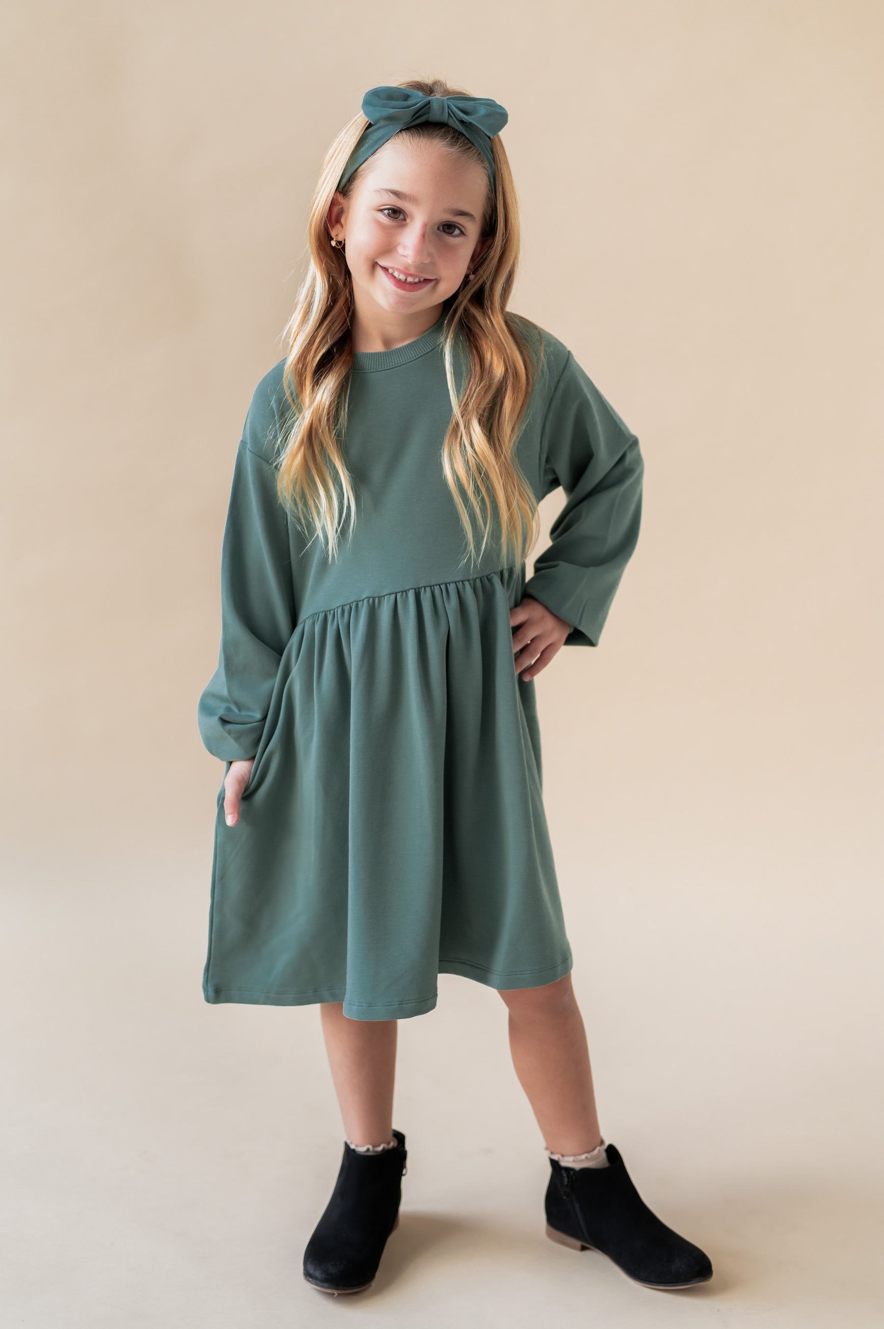 Girl posing in Long Sleeve Skater Dress in Pine color with similar color knotted bow headband on a beige background