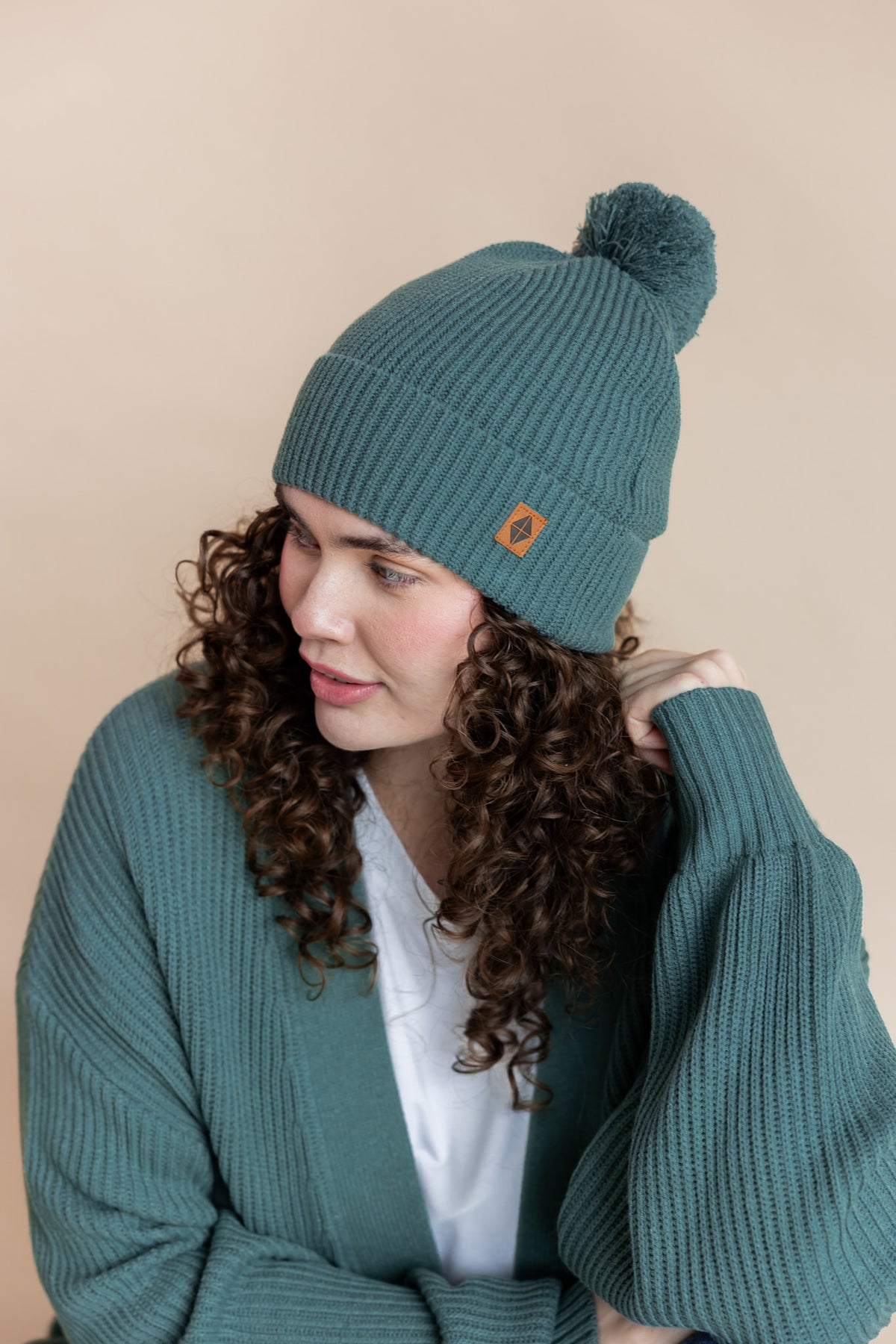 Modal wearing Chunky Knit Adult Pom in Pine over white T-shirt and similar color chunky knit cardigan on beige background