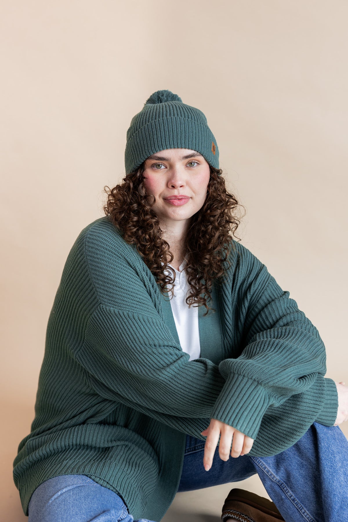 Modal wearing Chunky Knit Adult Pom in Pine over white T-shirt, similar color chunky knit cardigan and blue jeans on beige background