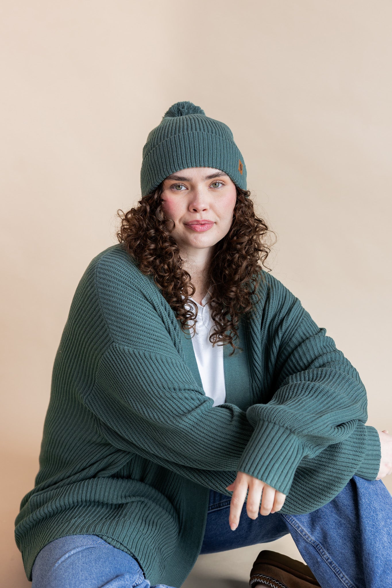 Modal wearing Chunky Knit Adult Pom in Pine over white T-shirt, similar color chunky knit cardigan and blue jeans on beige background