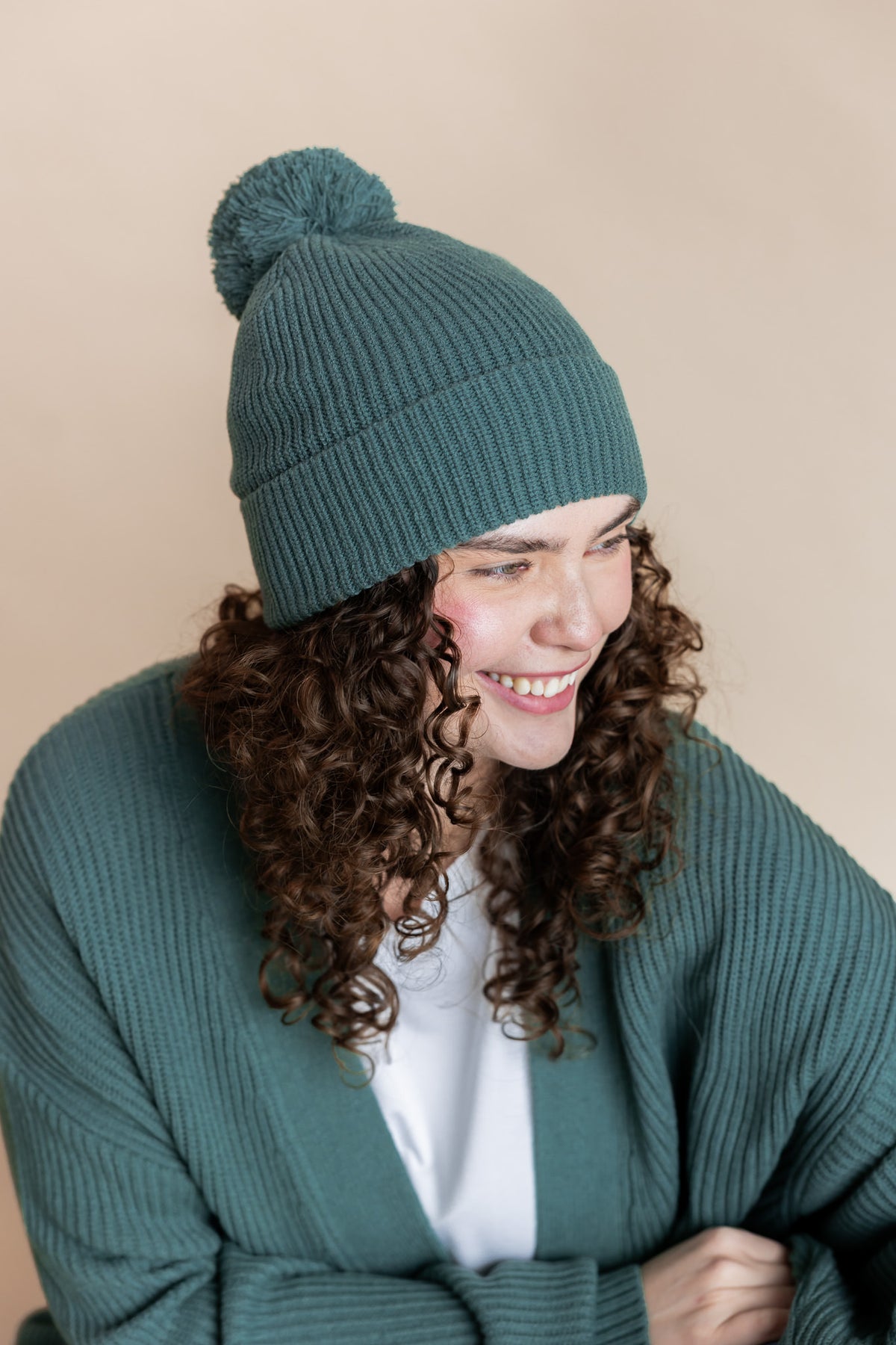 Modal smiling in Chunky Knit Adult Pom in Pine over white T-shirt and similar color chunky knit cardigan on beige background