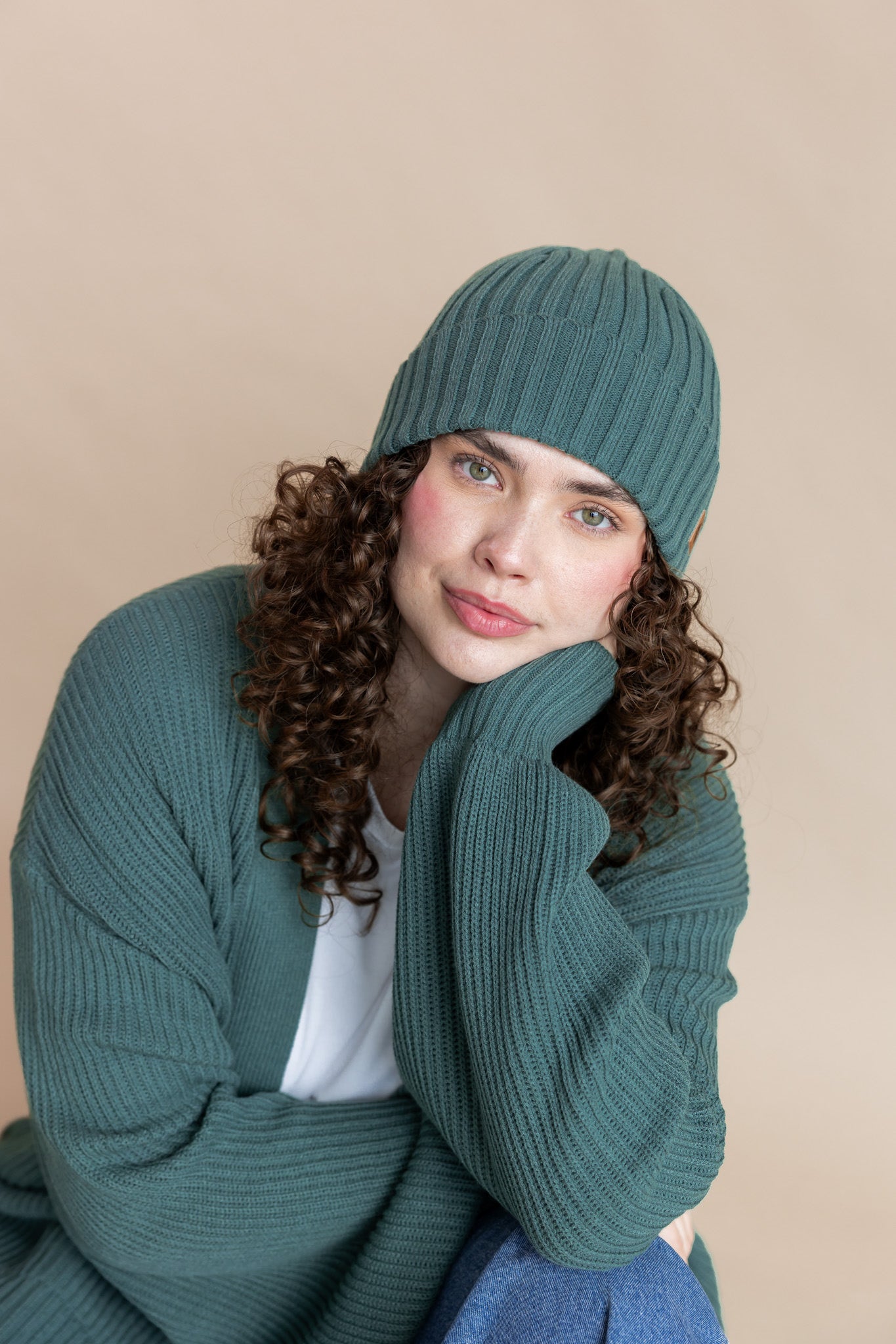Modal wearing Chunky Knit Adult Ribbed Beanie in Pine over white T-shirt and Chunky knit cardigan in Pine on a beige background