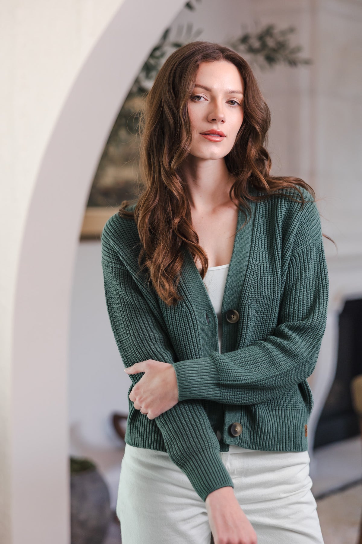 Modal standing in living room while posing in Chunky Knit Women's Cropped Cardigan in Pine over white pants