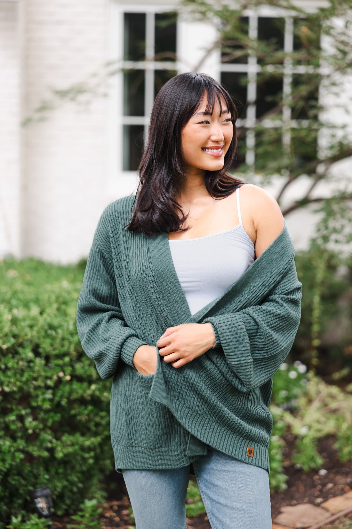 Modal smiling outdoors wearing Chunky knit oversized cardigan in pine over white camisole and blue jeans