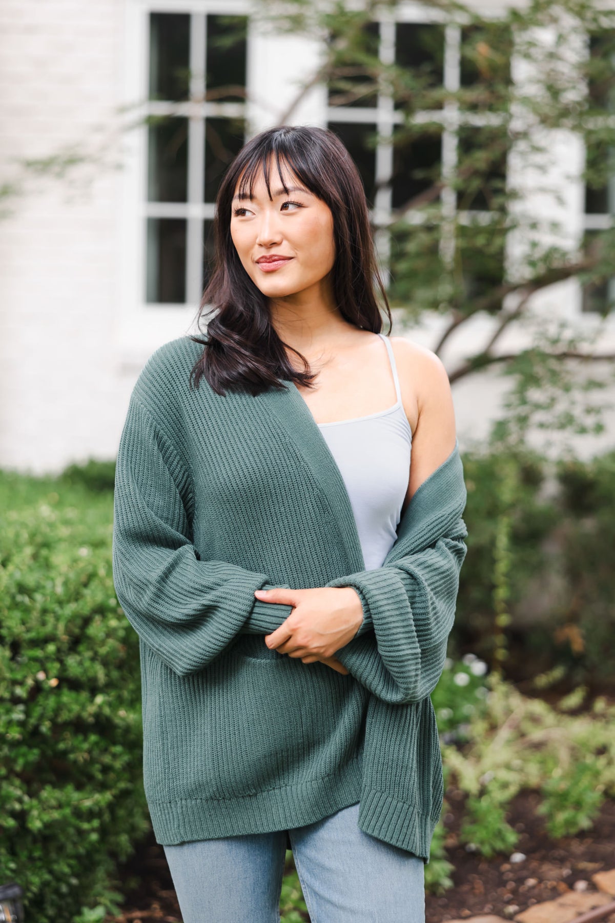 Modal standing outdoors wearing Chunky knit oversized cardigan in pine over white camisole and blue jeans