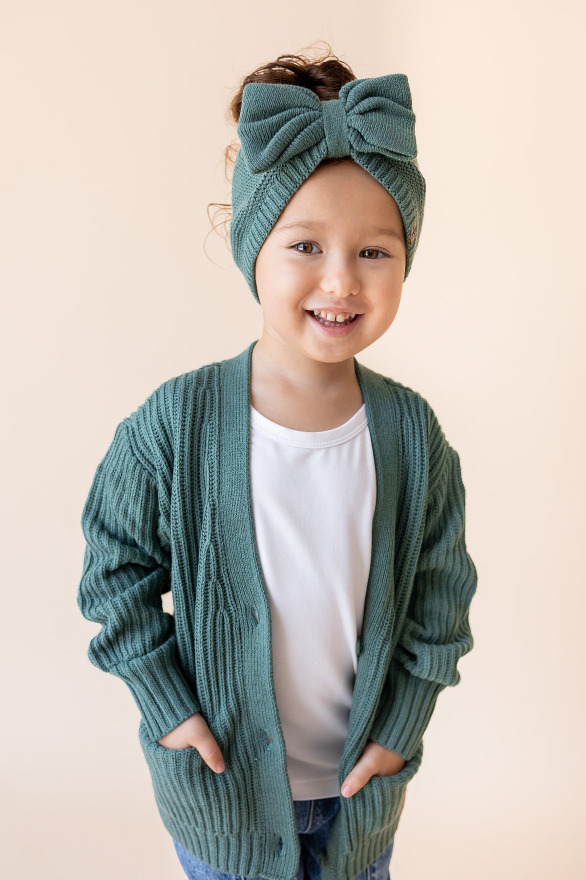 Child wearing Chunky Knit Oversized Cardigan in Pine over white t-shirt, blue jeans and chunky knit bow headband in pine on a beige background