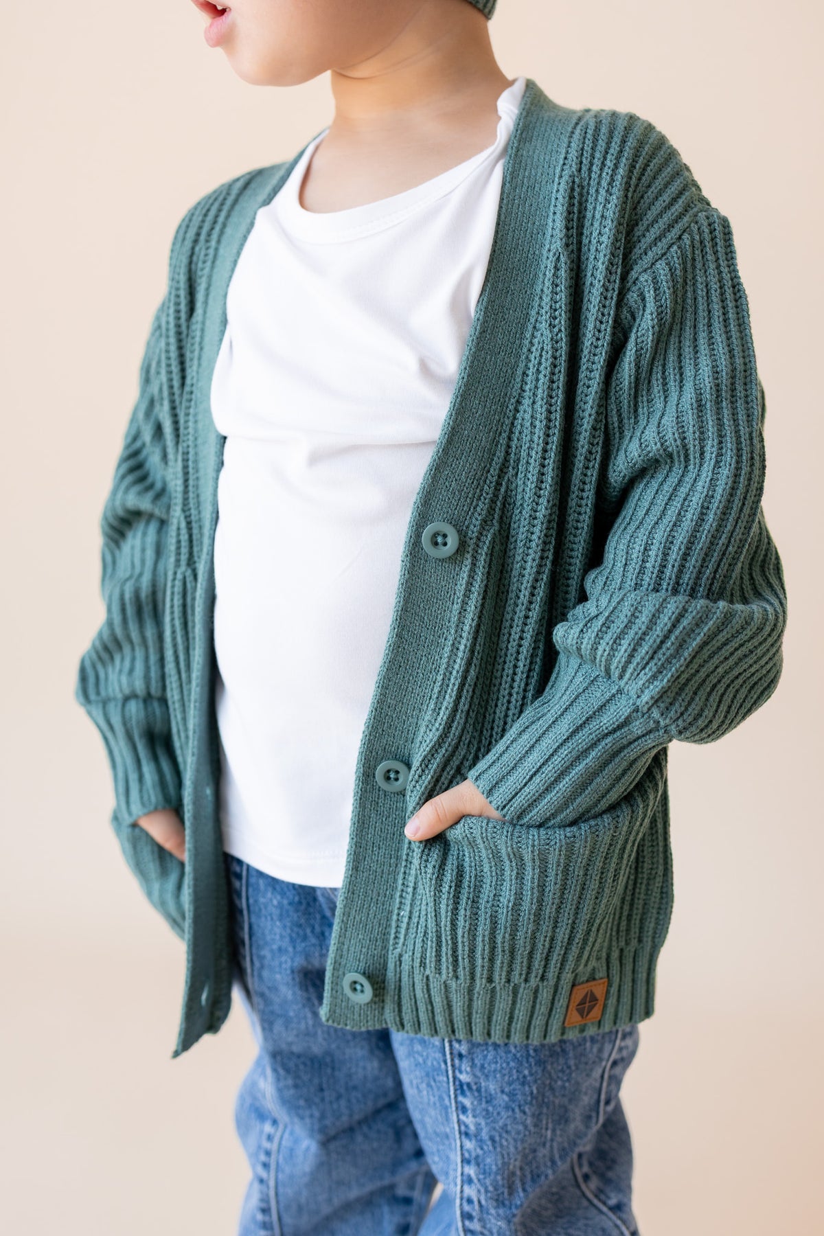 Close up of Child wearing Chunky Knit Oversized Cardigan in Pine over white t-shirt, blue jeans on a beige background