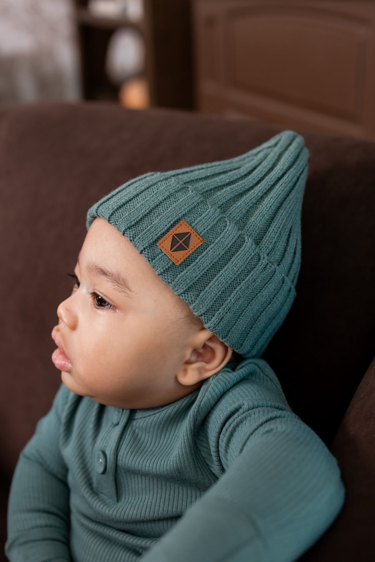 Baby wearing Chunky knit ribbed beanie in pine over pine romper on a brown sofa