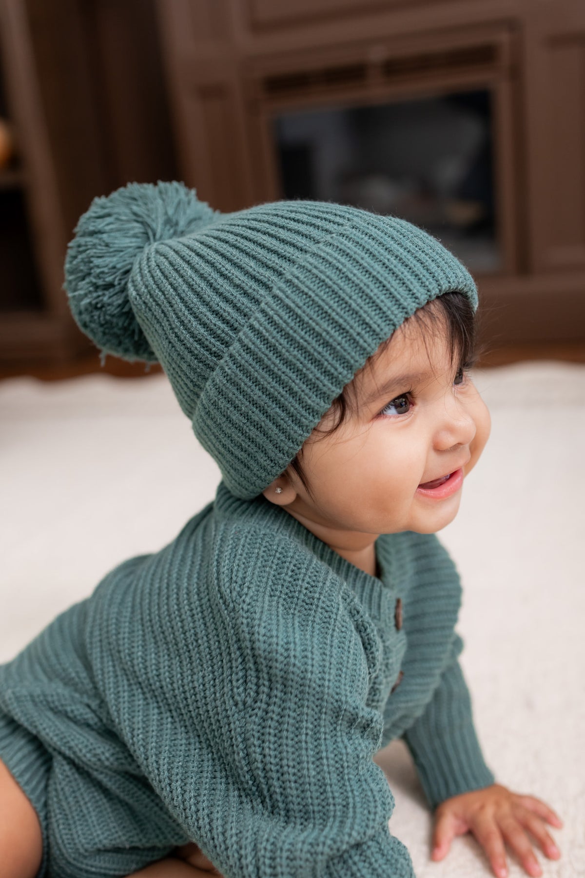 Baby wearing Chunky Knit Pom Beanie in Pine over chunky knit romper on white carpet