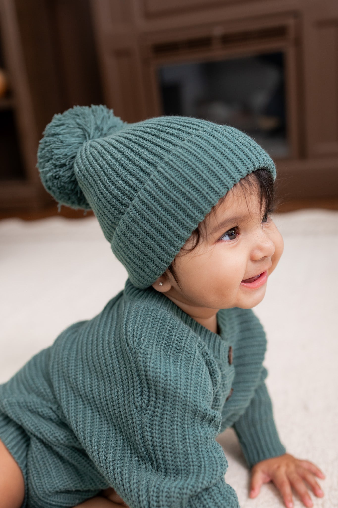 Baby wearing Chunky Knit Pom Beanie in Pine over chunky knit romper on white carpet