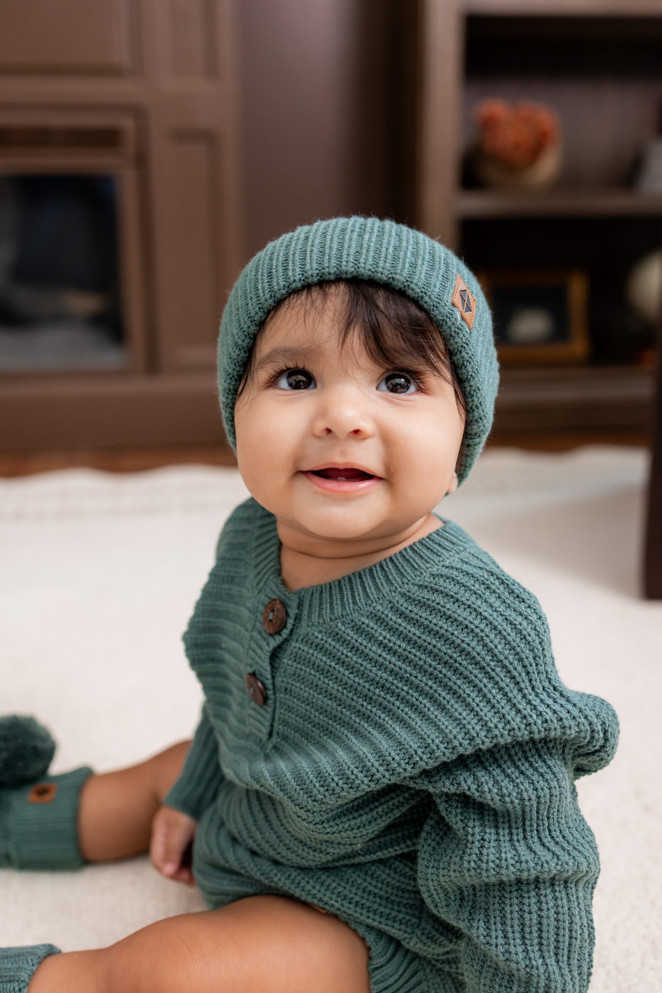 Baby wearing Chunky Knit Romper and chunky knit pom beanie in Pine sitting on a white carpet