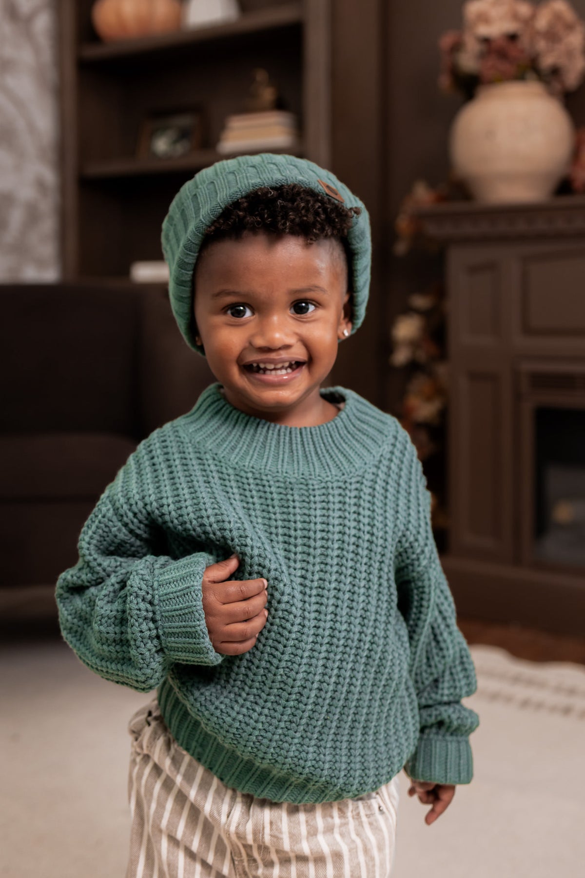 Boy wearing Chunky Knit Sweater and chunky knit ribbed beanie in Pine over beige pants standing in a living room