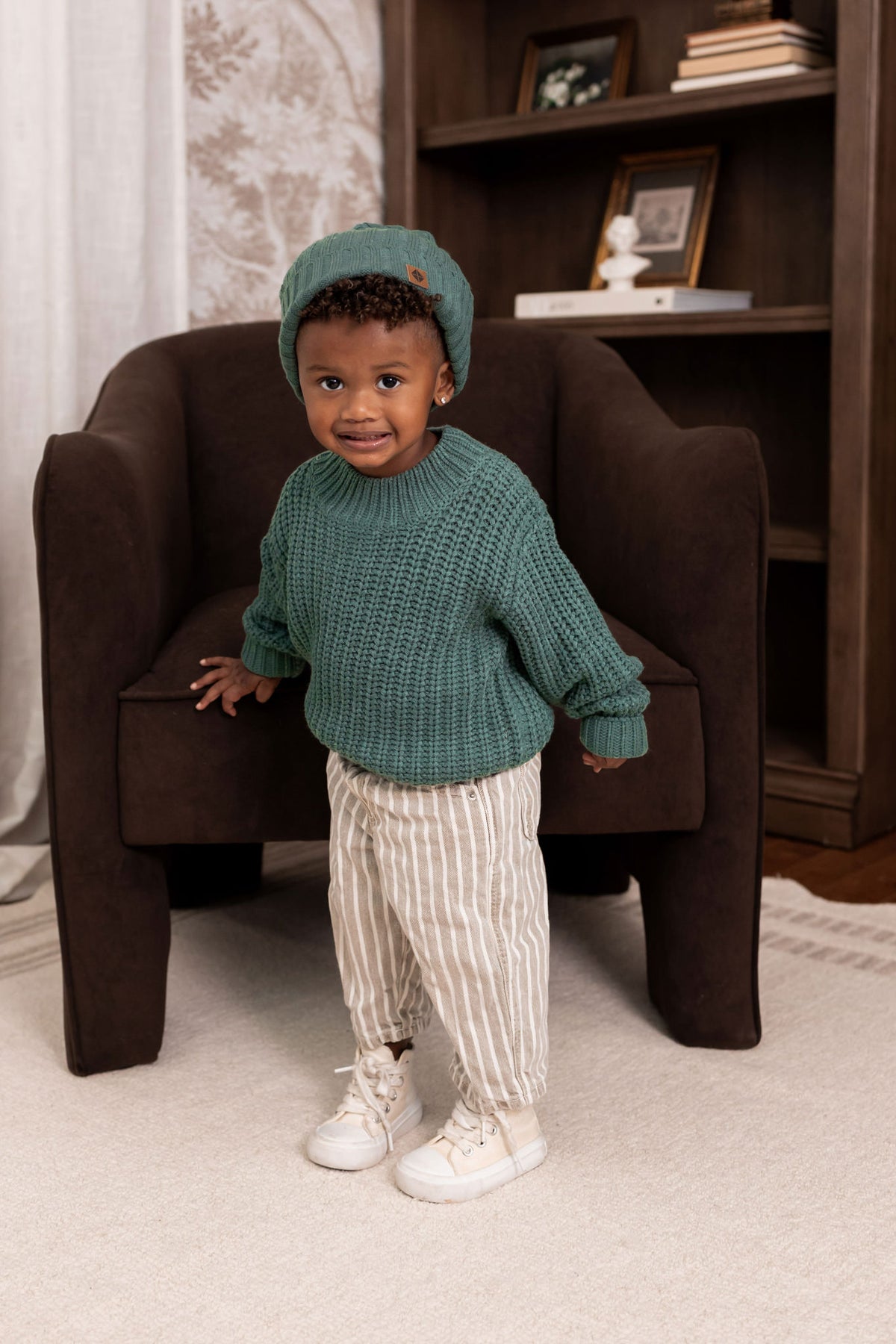 Boy wearing Chunky Knit Sweater and chunky knit ribbed beanie in Pine over beige pants standing in a living room